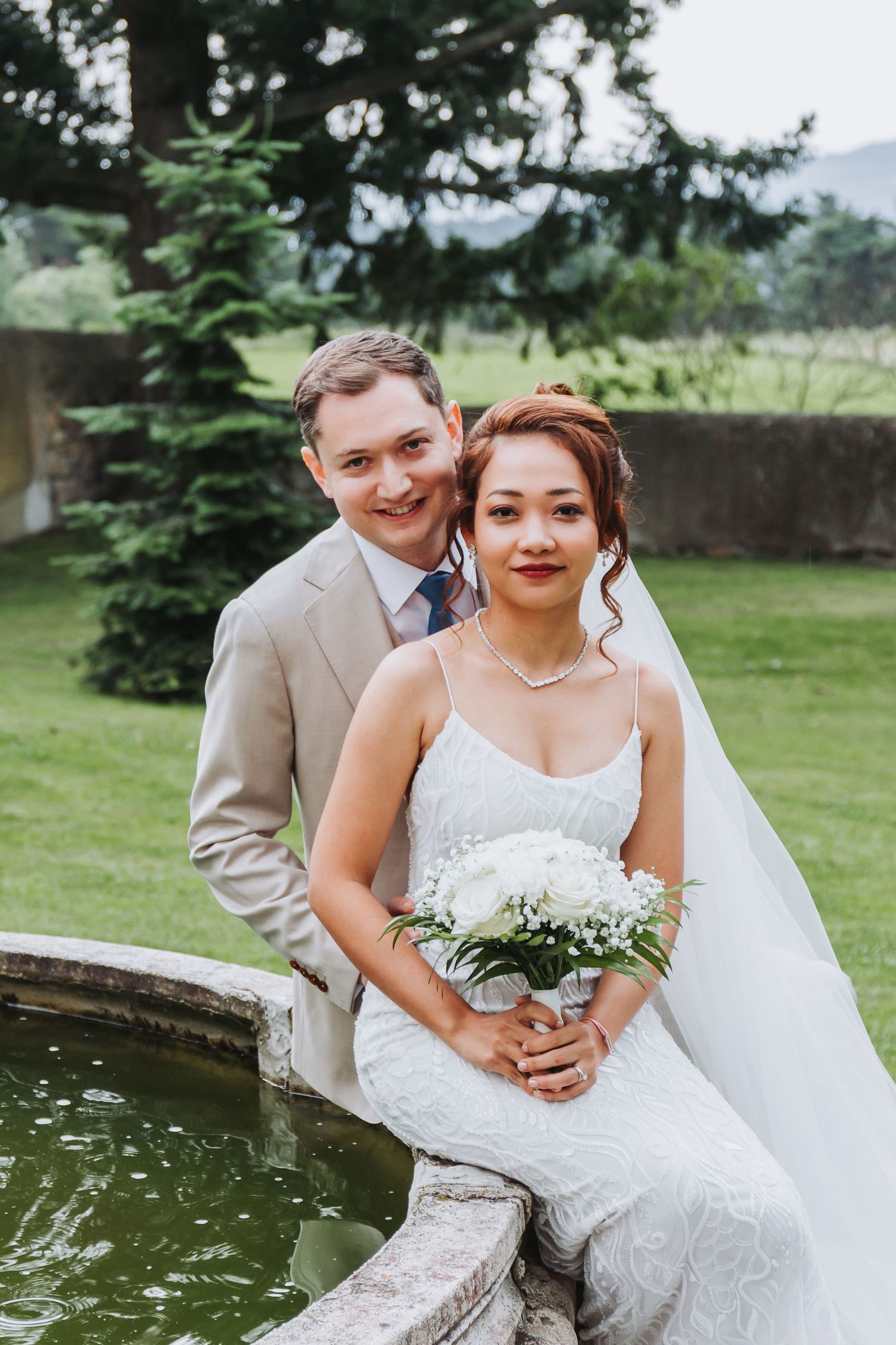 Bride in white lace gown holding bouquet sits with groom by a stone fountain in a lush green garden.