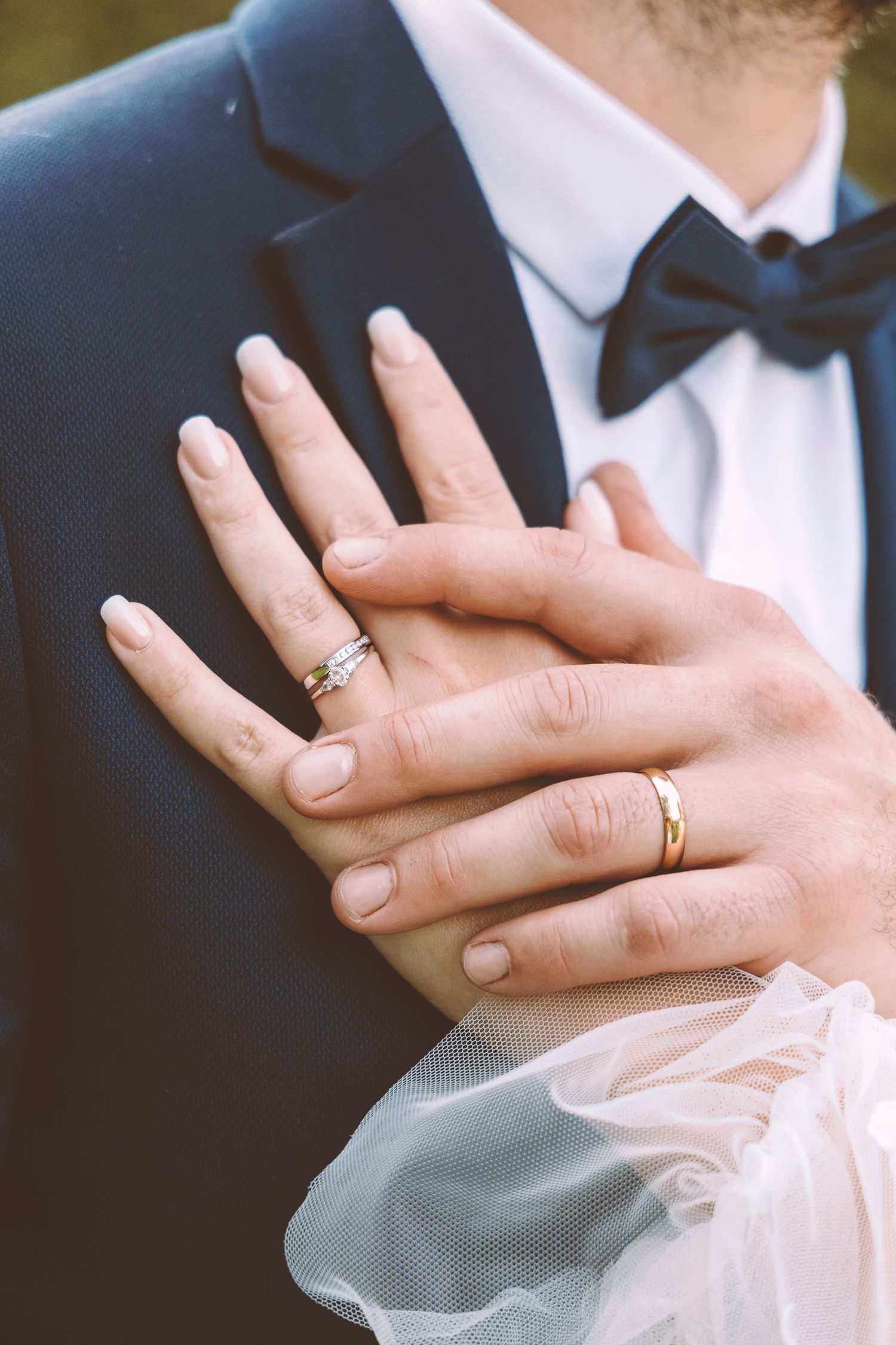 Close-up of bride and groom hands showing wedding rings, groom in navy suit with black bow tie.