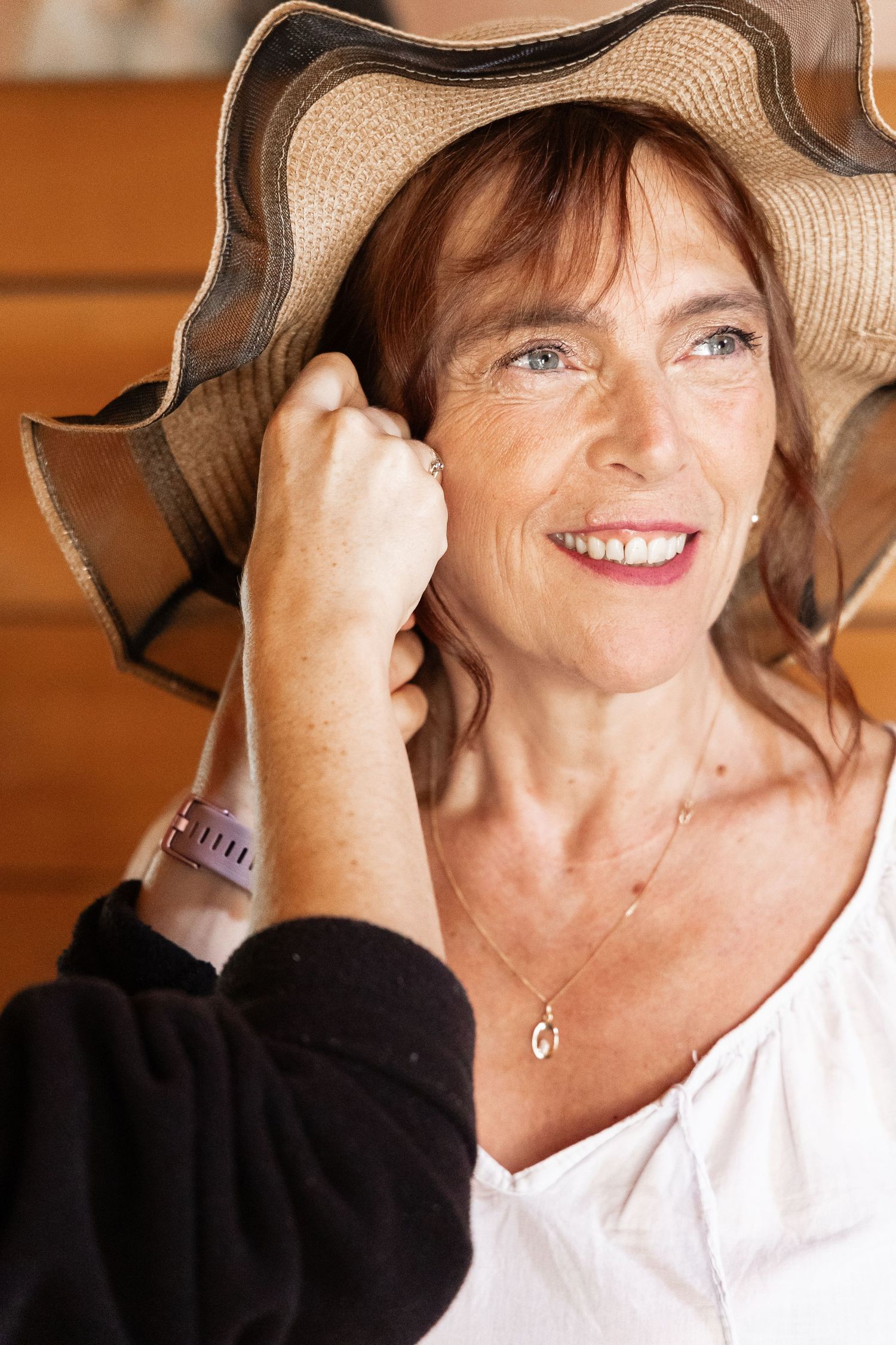 Elegant mature woman smiling while adjusting a wide-brim straw hat indoors.