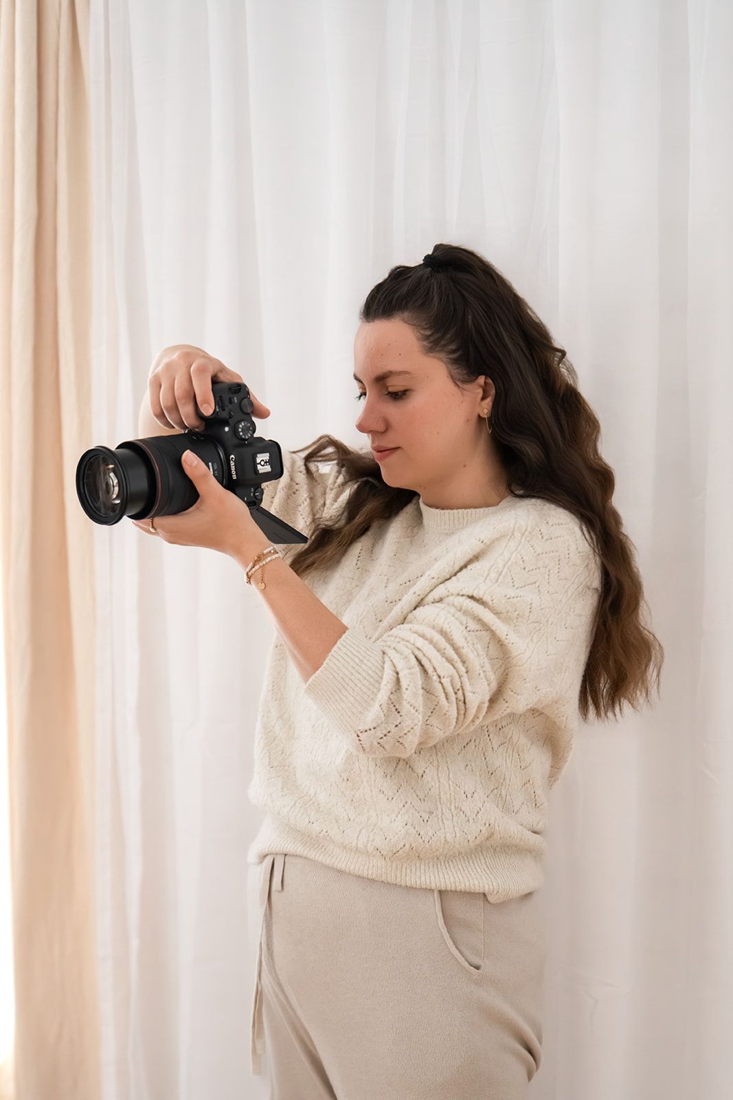 A young woman in a beige sweater reviews photos on her Canon DSLR camera against a white curtain backdrop.