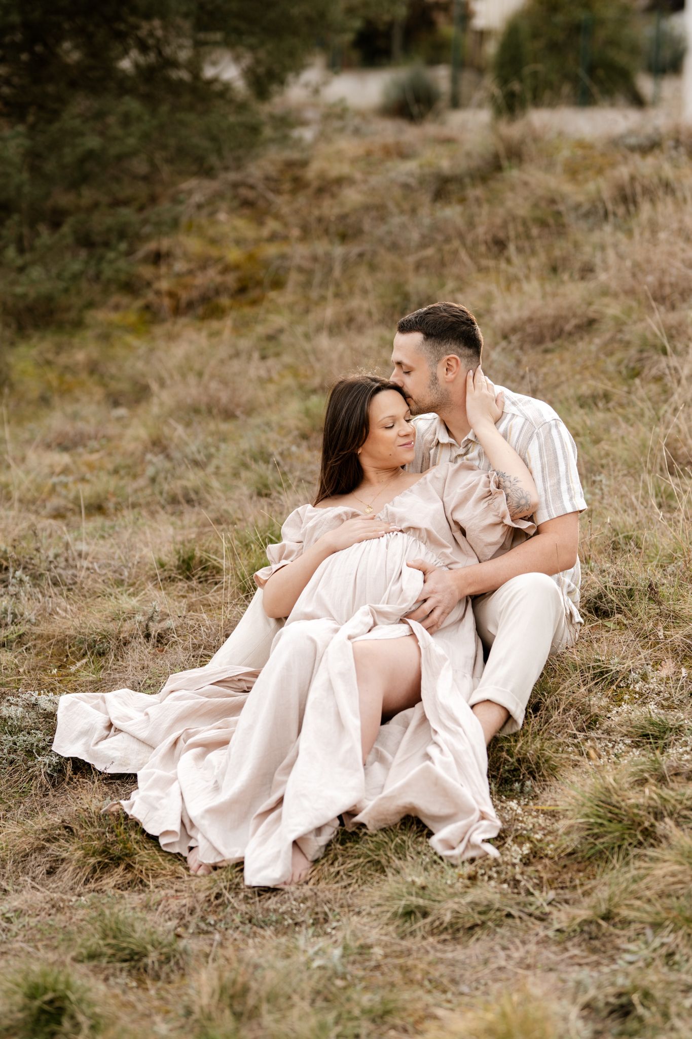 Photo de grossesse en Isère : la douceur d'un futur papa entourant sa femme en robe beige, assis dans l'herbe.