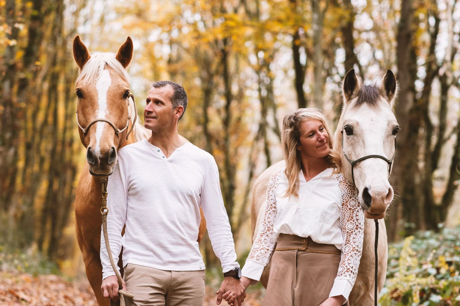 Couple holding hands walking with two horses along a golden autumn forest path.