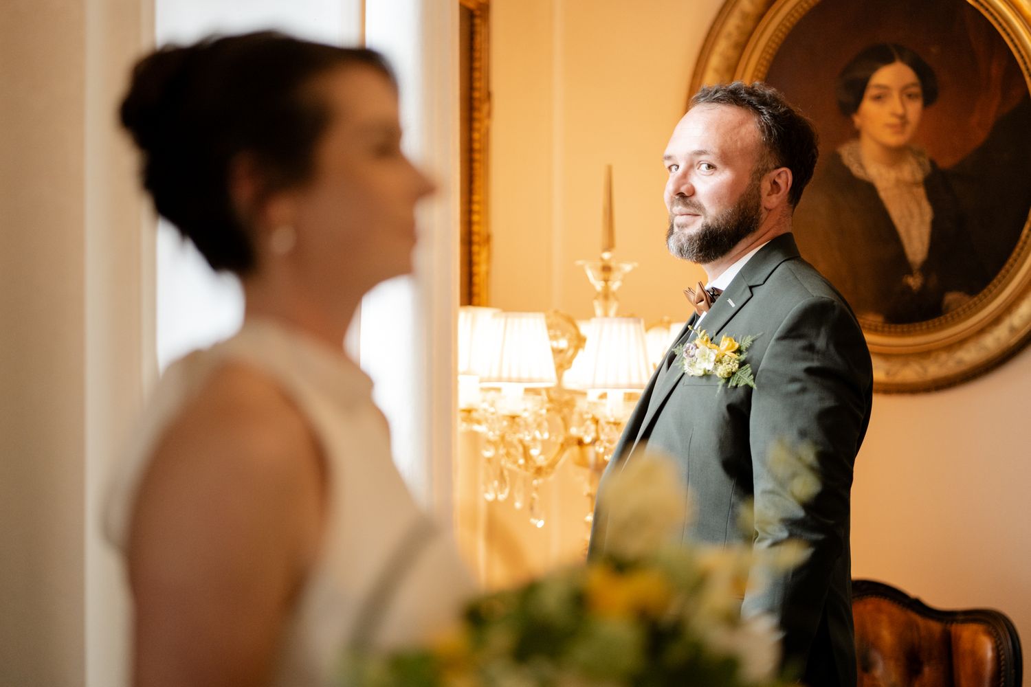 Groom gazing at bride in elegant room with chandelier and classical portrait on wall.