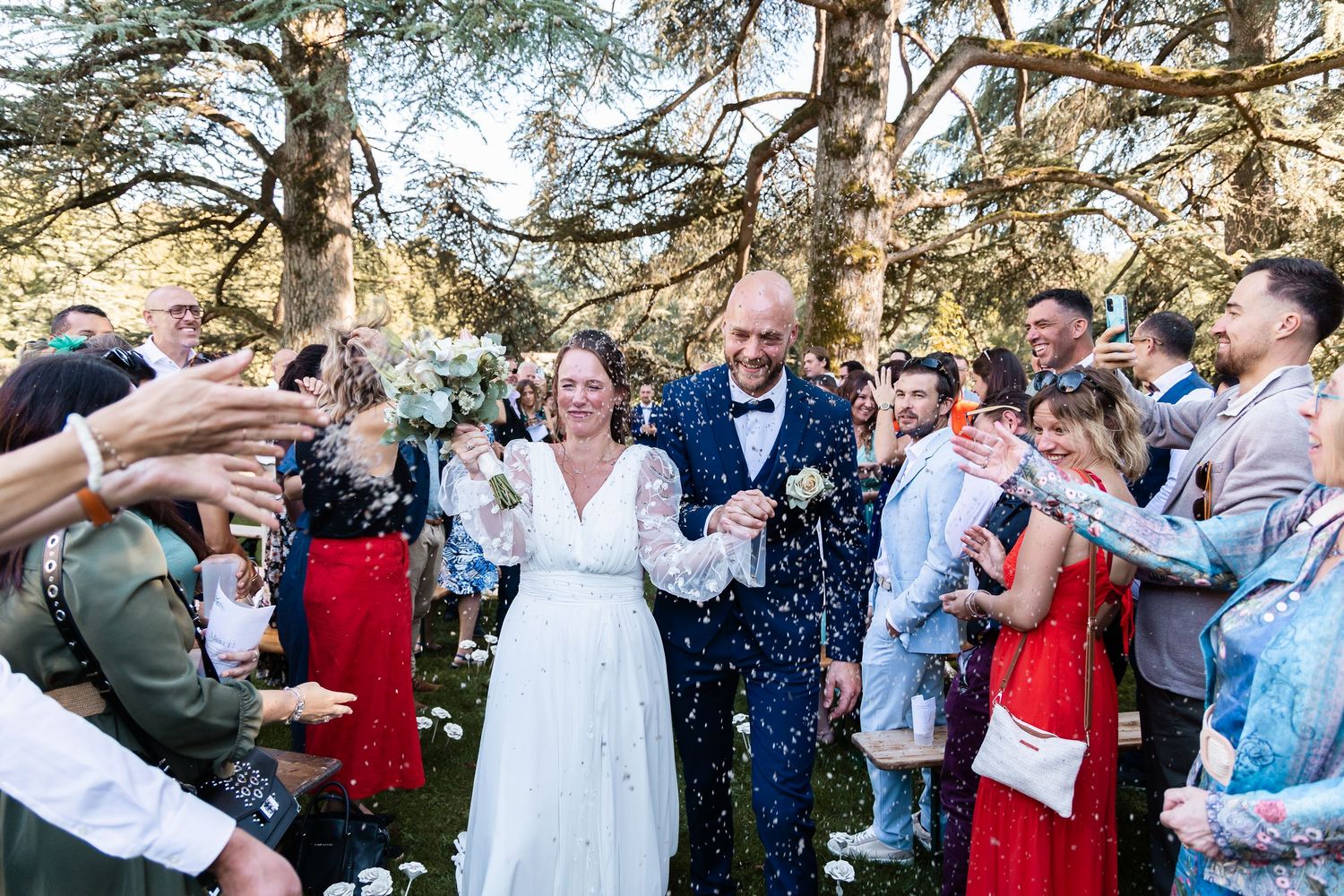 Happy newlyweds walk hand in hand through confetti as smiling guests celebrate their outdoor wedding.