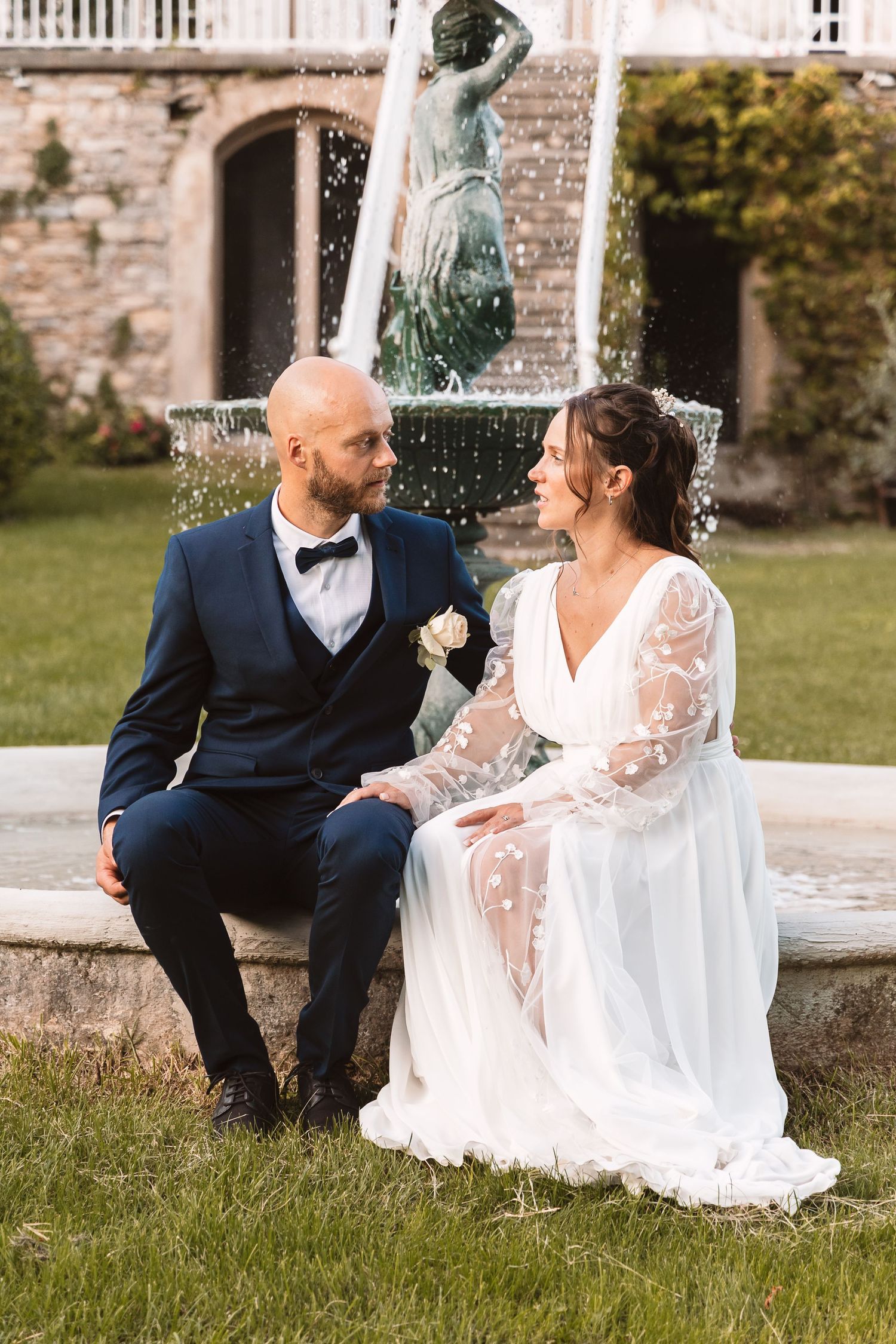 Bride and groom sit together by ornate stone fountain, smiling at each other in elegant garden setting.
