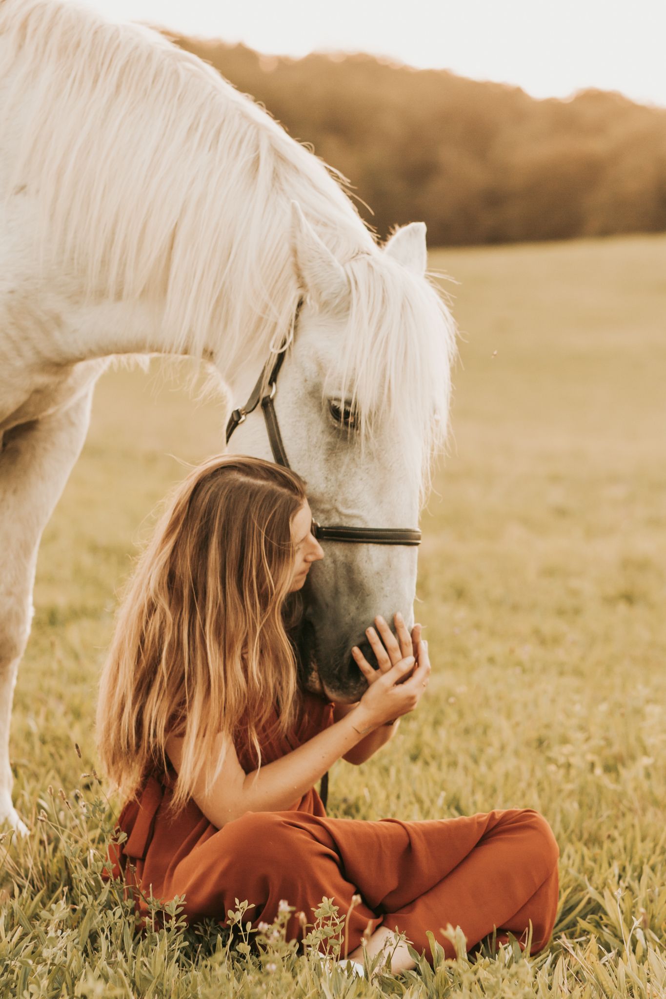 A woman in rust outfit sits cross-legged feeding a white horse in a meadow.