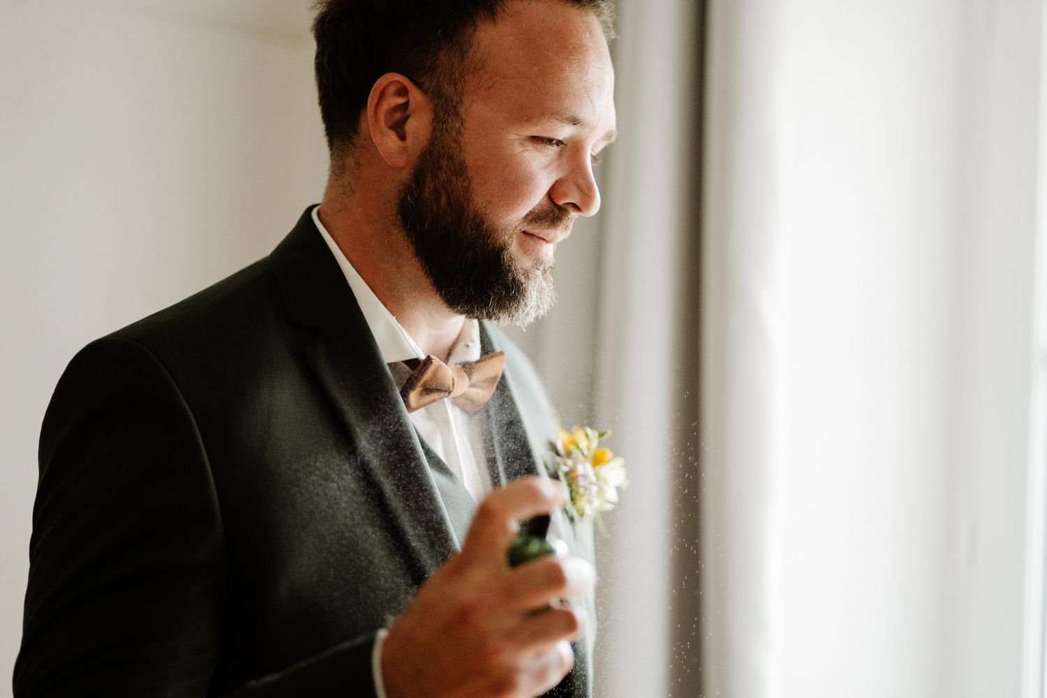 Bearded groom in dark green suit admiring boutonniere by window while preparing for wedding day.