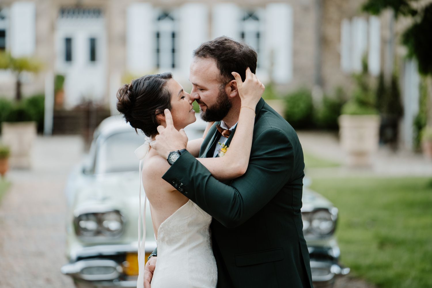 Bride and groom sharing a tender moment, almost kissing, with a vintage car and chateau in the background.