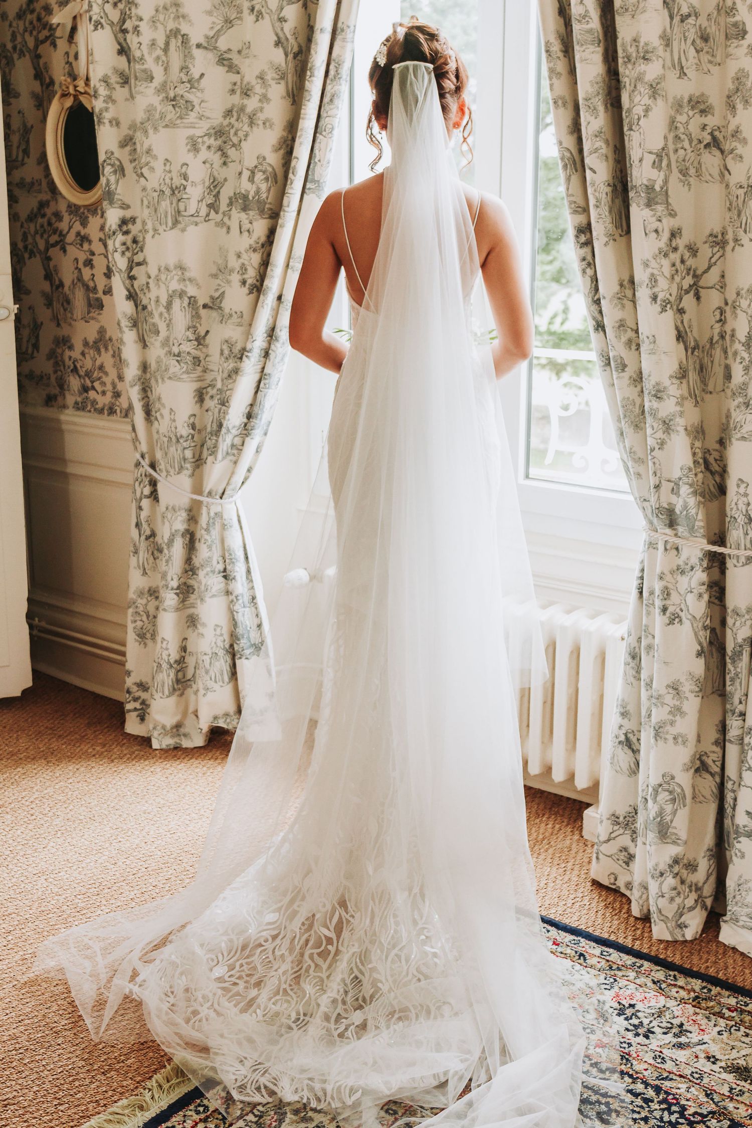 Bride in white lace gown with long cathedral veil standing at window with floral toile curtains.