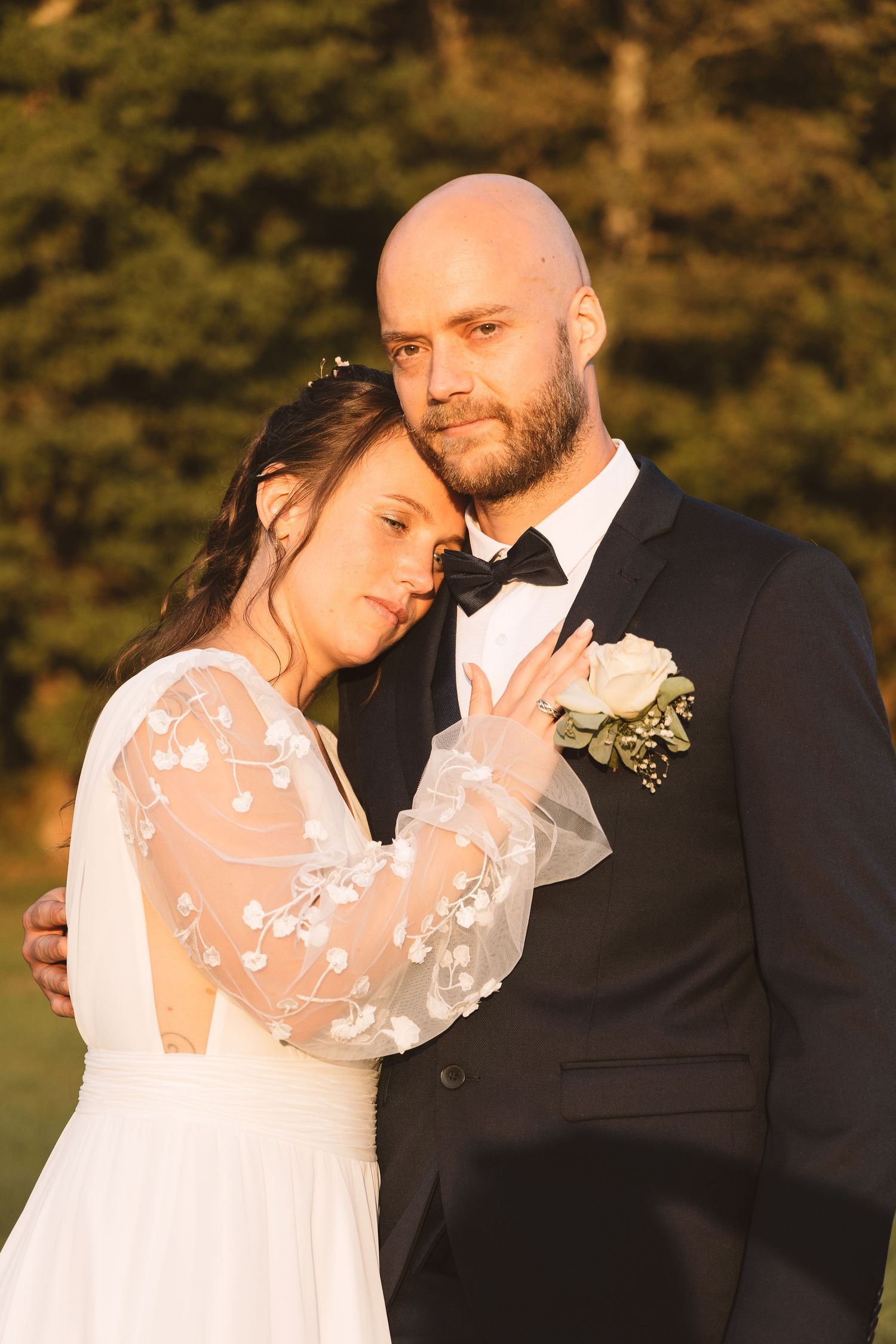 Bride in lace wedding dress rests head on groom's chest as couple embraces in warm golden hour light.