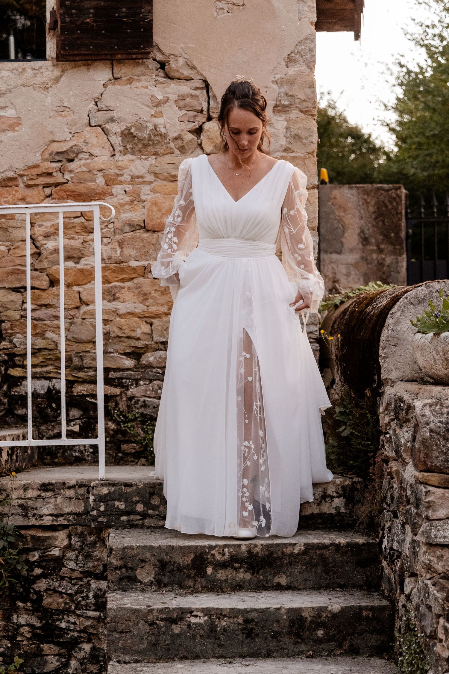 Elegant bride in white V-neck long sleeve floral wedding gown descending rustic stone steps outdoors.