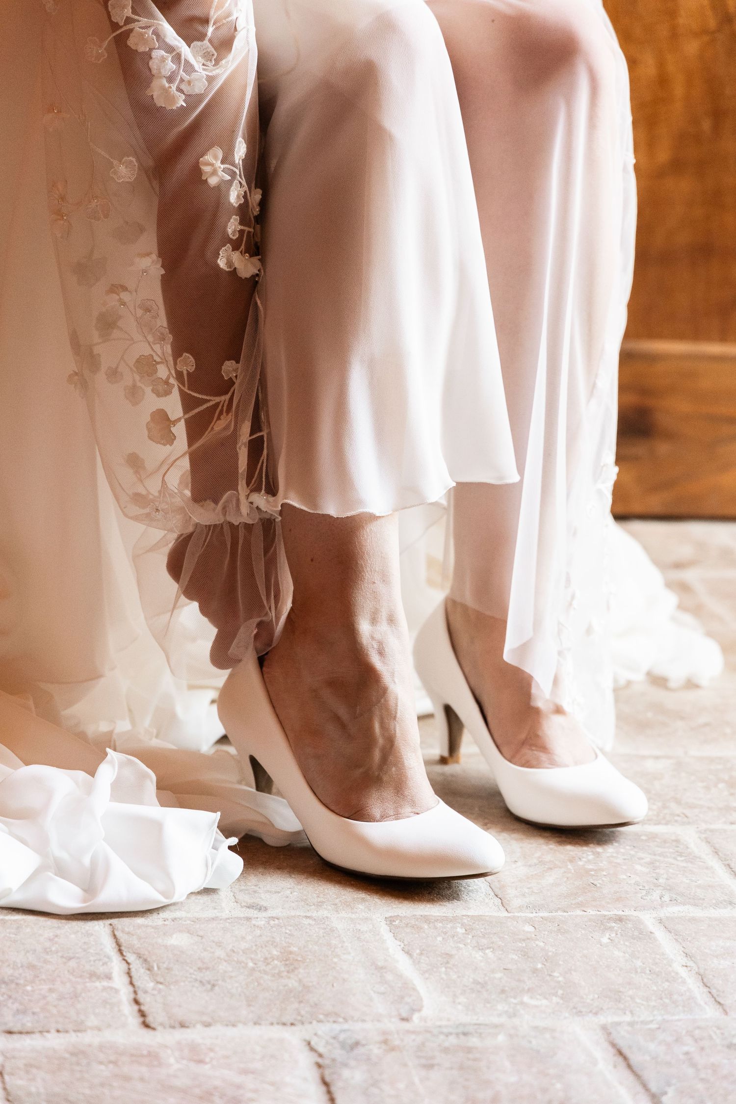 Close-up of bride's white satin heels beneath floral embroidered wedding dress hem.