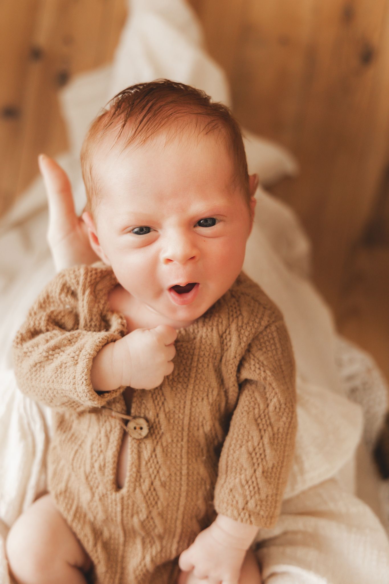 Adorable newborn baby in knit brown onesie with mouth open and hand raised looking at camera.