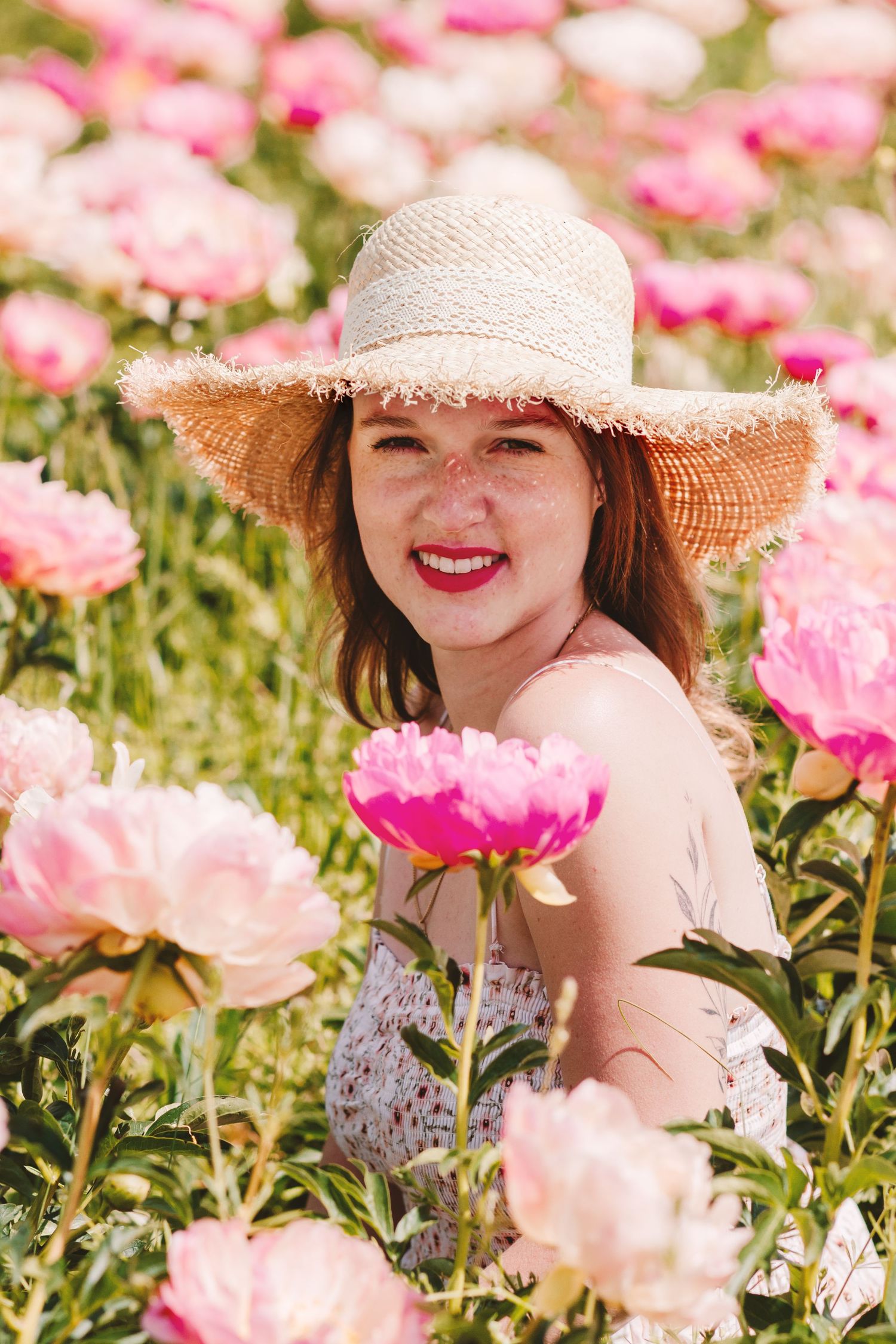 Smiling redhead woman in straw hat crouching among pink peonies in sunny field.