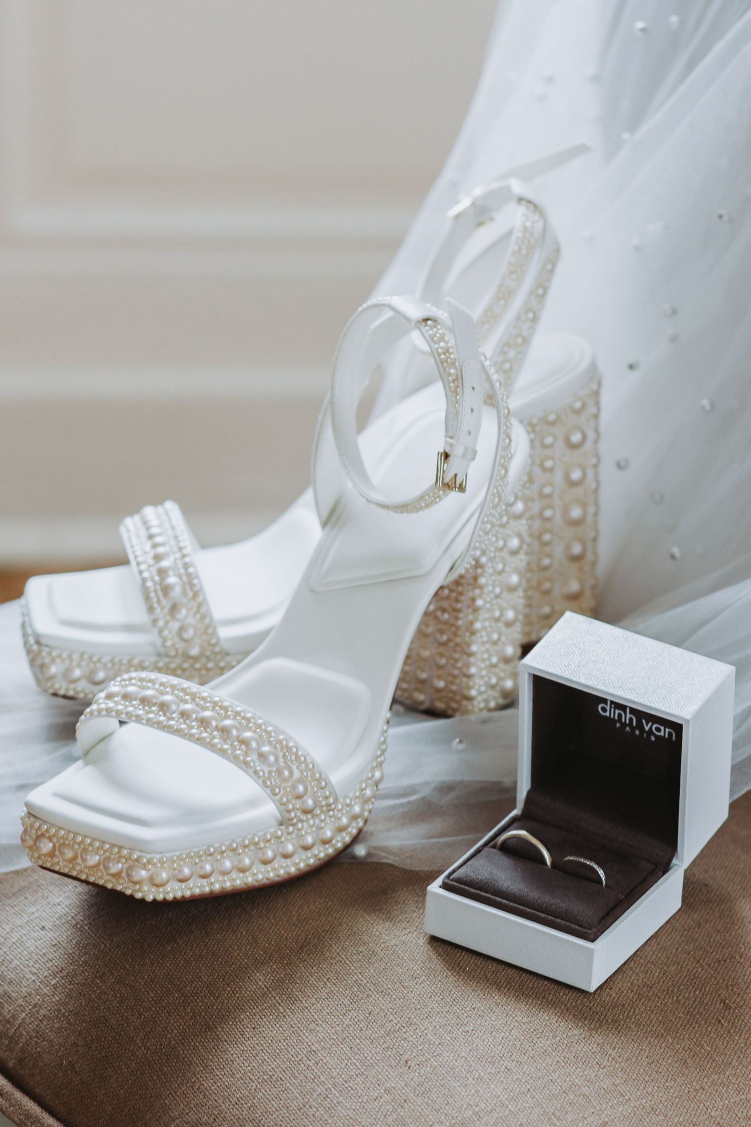 Pearl-embellished white bridal platform heels beside a Dinh Van ring box on a beige chair.