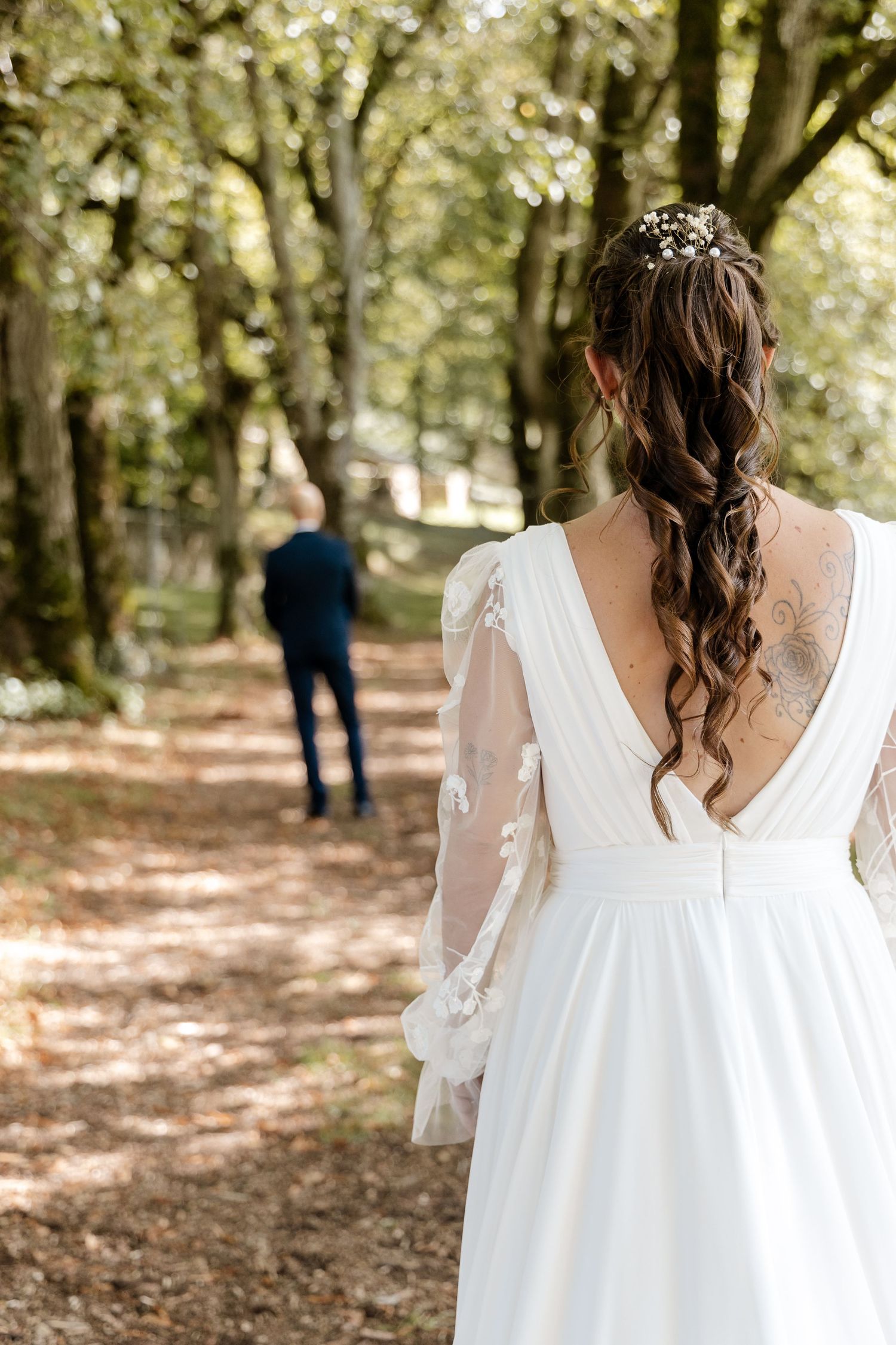 Bride in white lace gown with tattoo and floral hair piece walks toward groom in forest path.