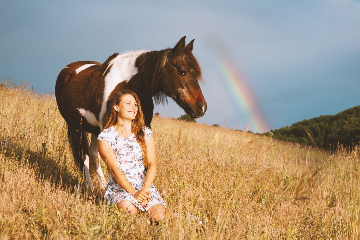 Smiling woman sitting in golden field beside a paint horse with rainbow behind.