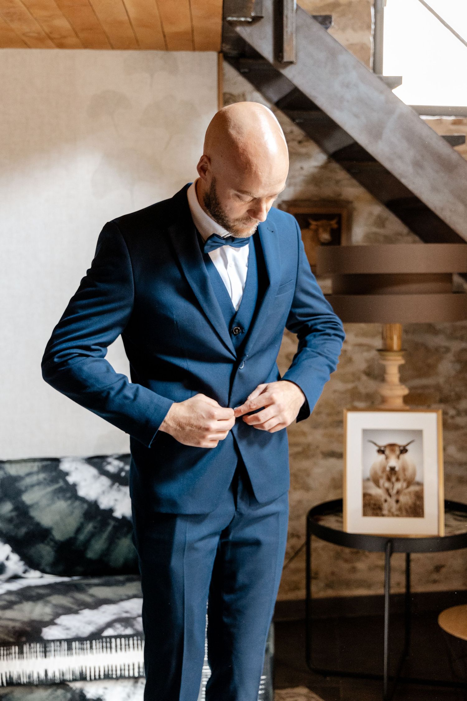 Groom in a navy blue three-piece suit buttoning his jacket near a staircase indoors.