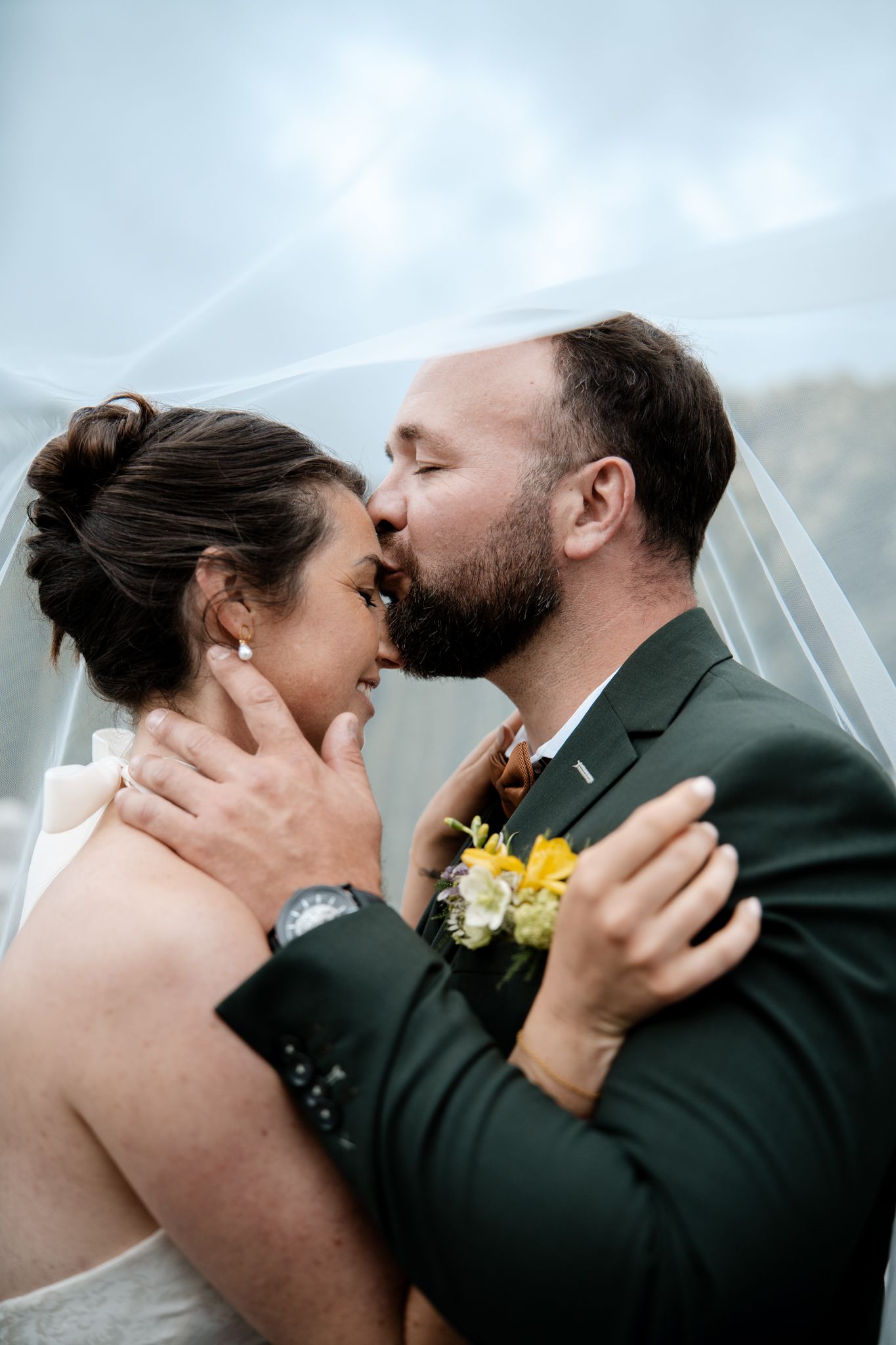 Groom with beard kissing bride on the cheek under a white veil, both embracing with yellow boutonniere.