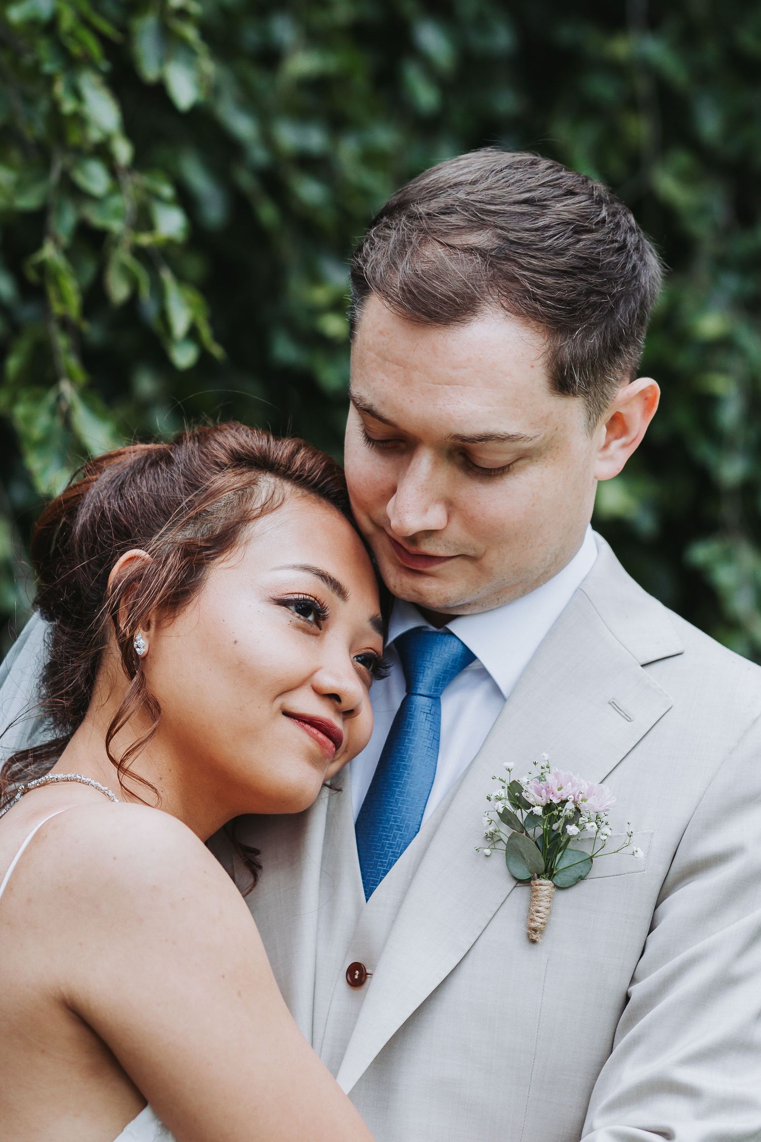 Close-up of bride leaning on groom's shoulder, groom in beige suit with pink boutonniere against green foliage.