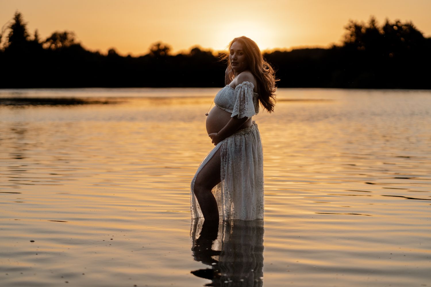 Pregnant woman in flowing white dress standing in lake water at stunning golden sunset, holding baby bump.