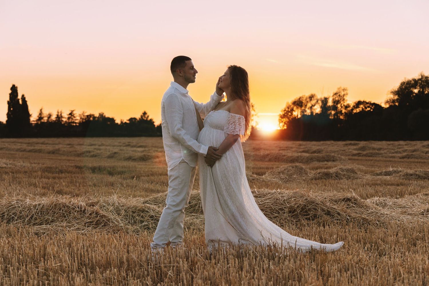 A couple embraces in a harvested field at sunset, the woman wearing a flowing white maternity gown.