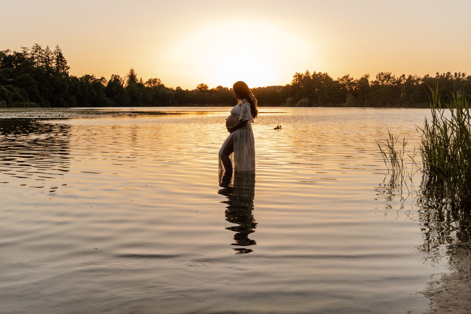 Pregnant woman in white dress standing alone in lake water at breathtaking golden sunset.