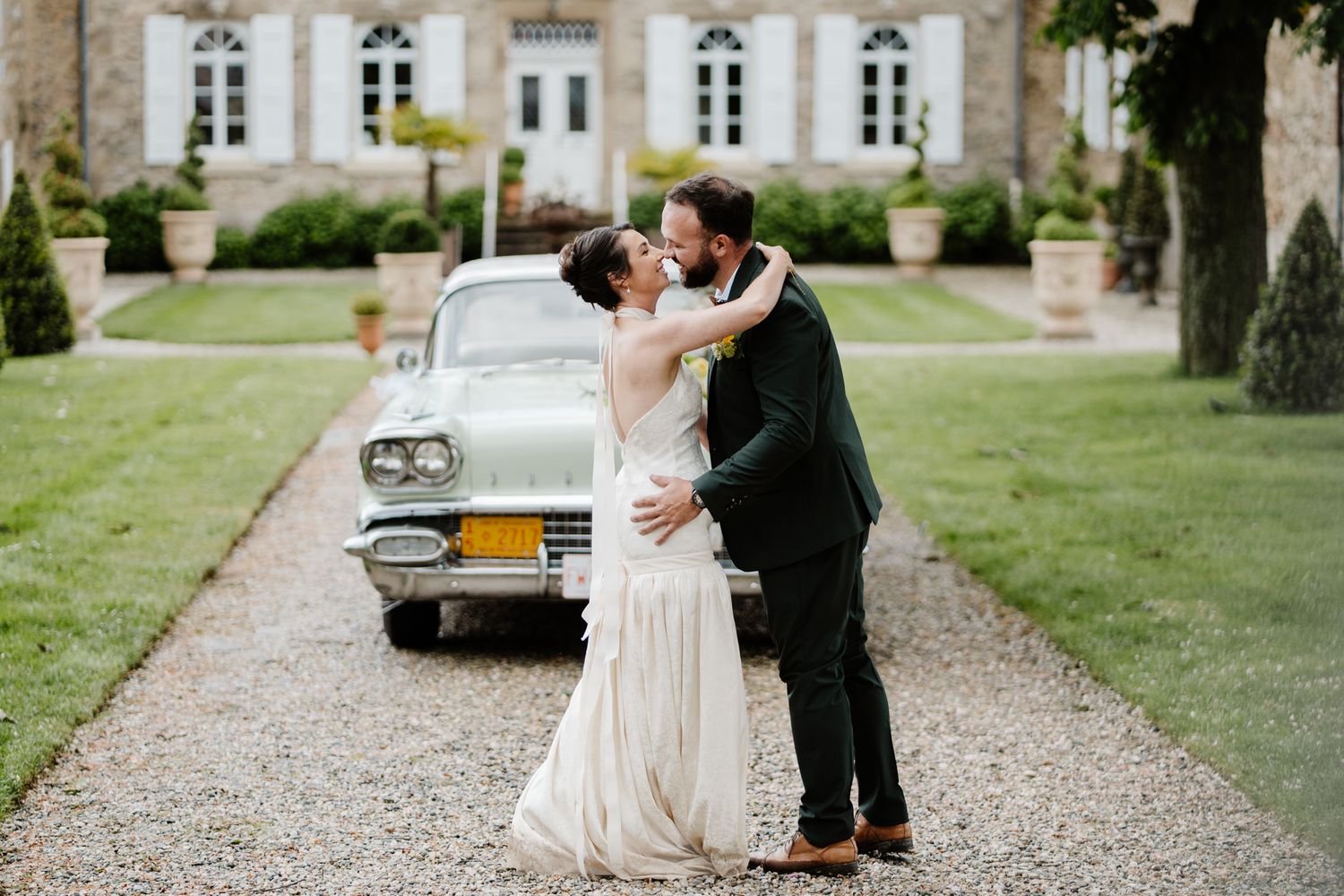 Bride and groom sharing romantic kiss beside vintage mint green car at chateau wedding venue.
