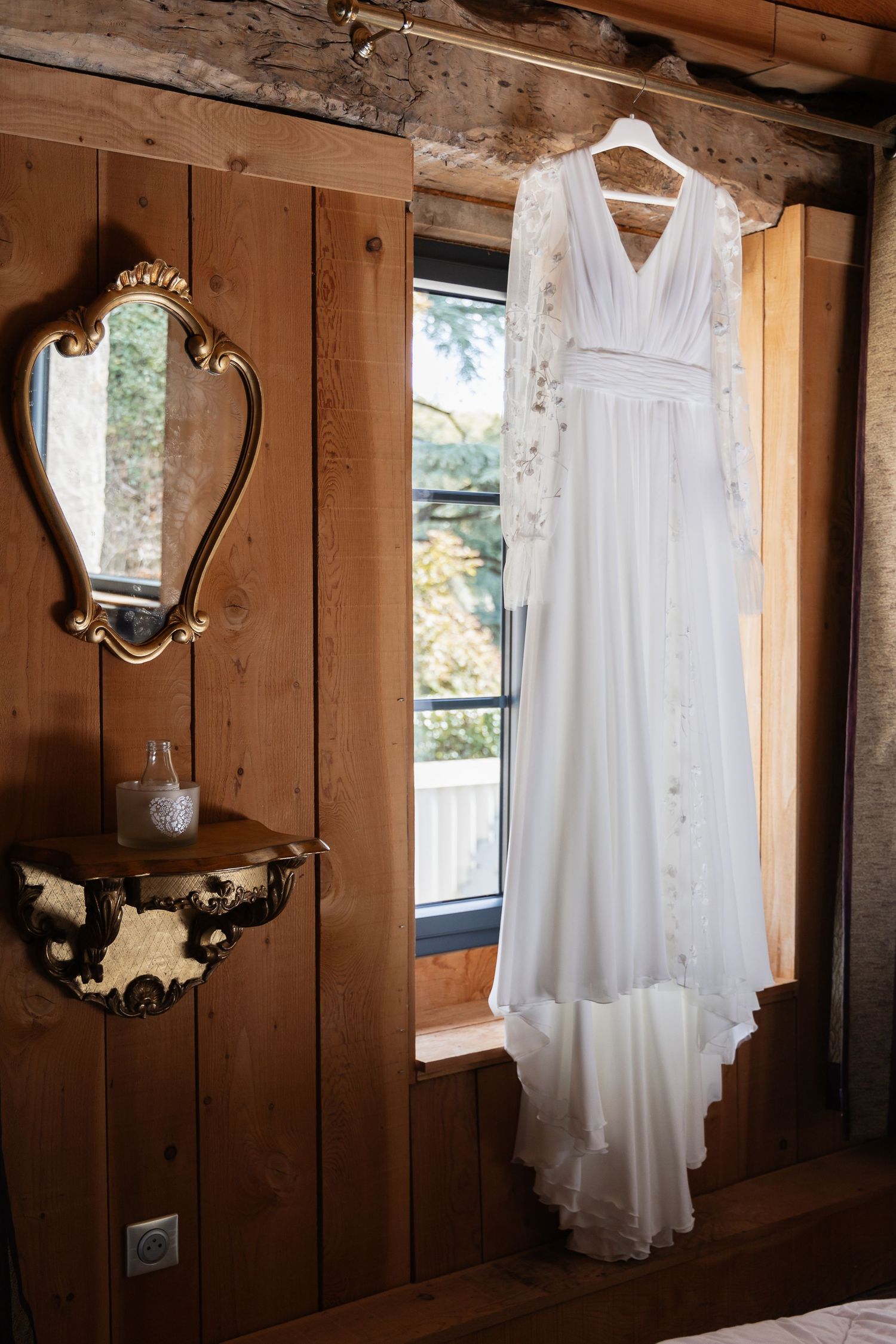 Elegant white long-sleeve floral wedding dress hanging on hanger by rustic wooden cabin window.