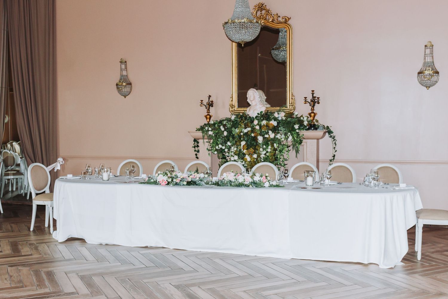 Elegant wedding head table decorated with white linen, floral garland, and candelabras in a ballroom.