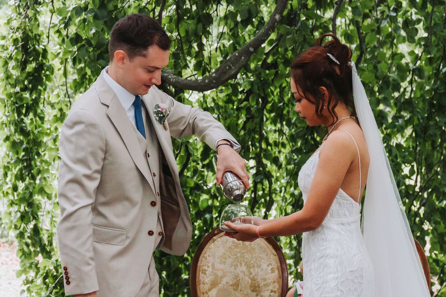 Groom in beige suit and bride in white lace dress perform a wedding ritual at an ornate chair under green foliage.