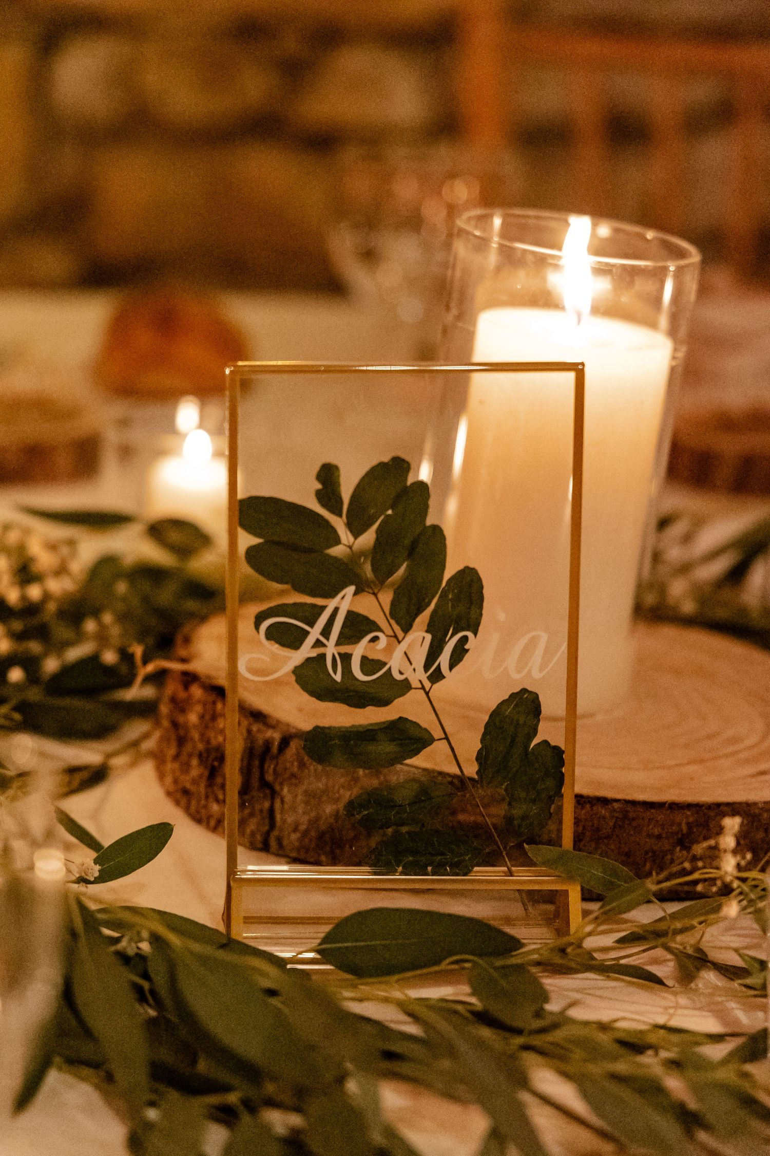 Gold-framed table sign with pressed leaves reading Acacia surrounded by candles and greenery at wedding.