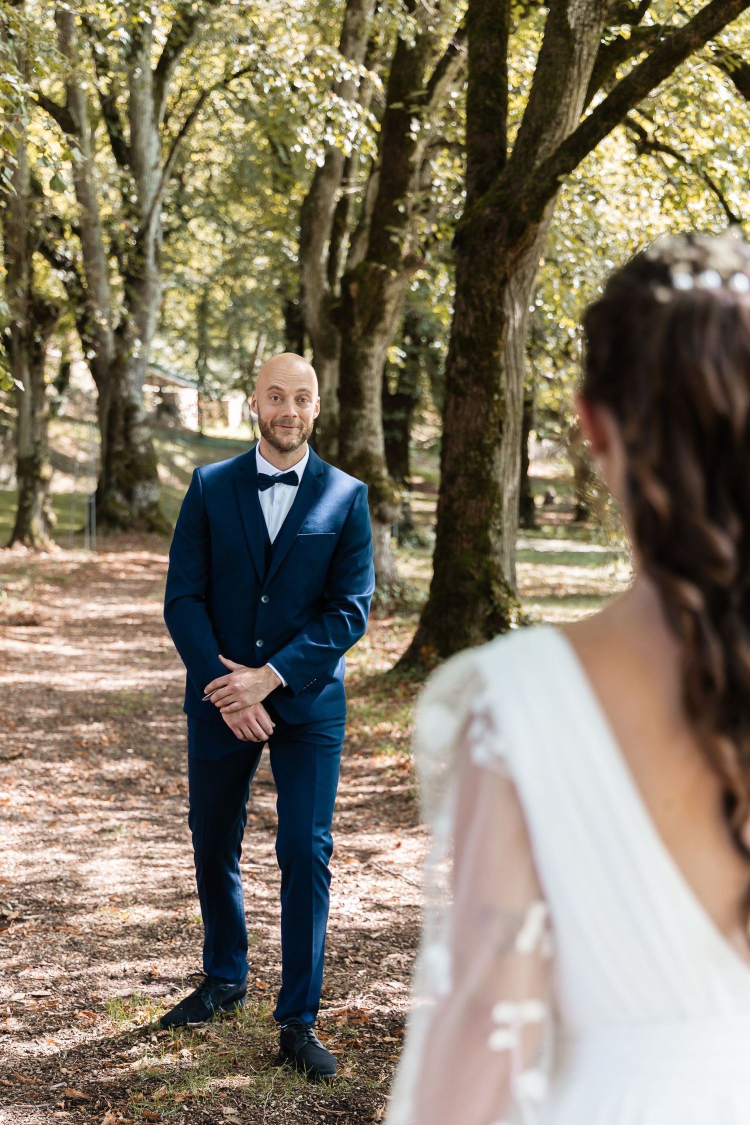Groom in navy tuxedo gazes at bride in embroidered white gown during first look moment in wooded setting.