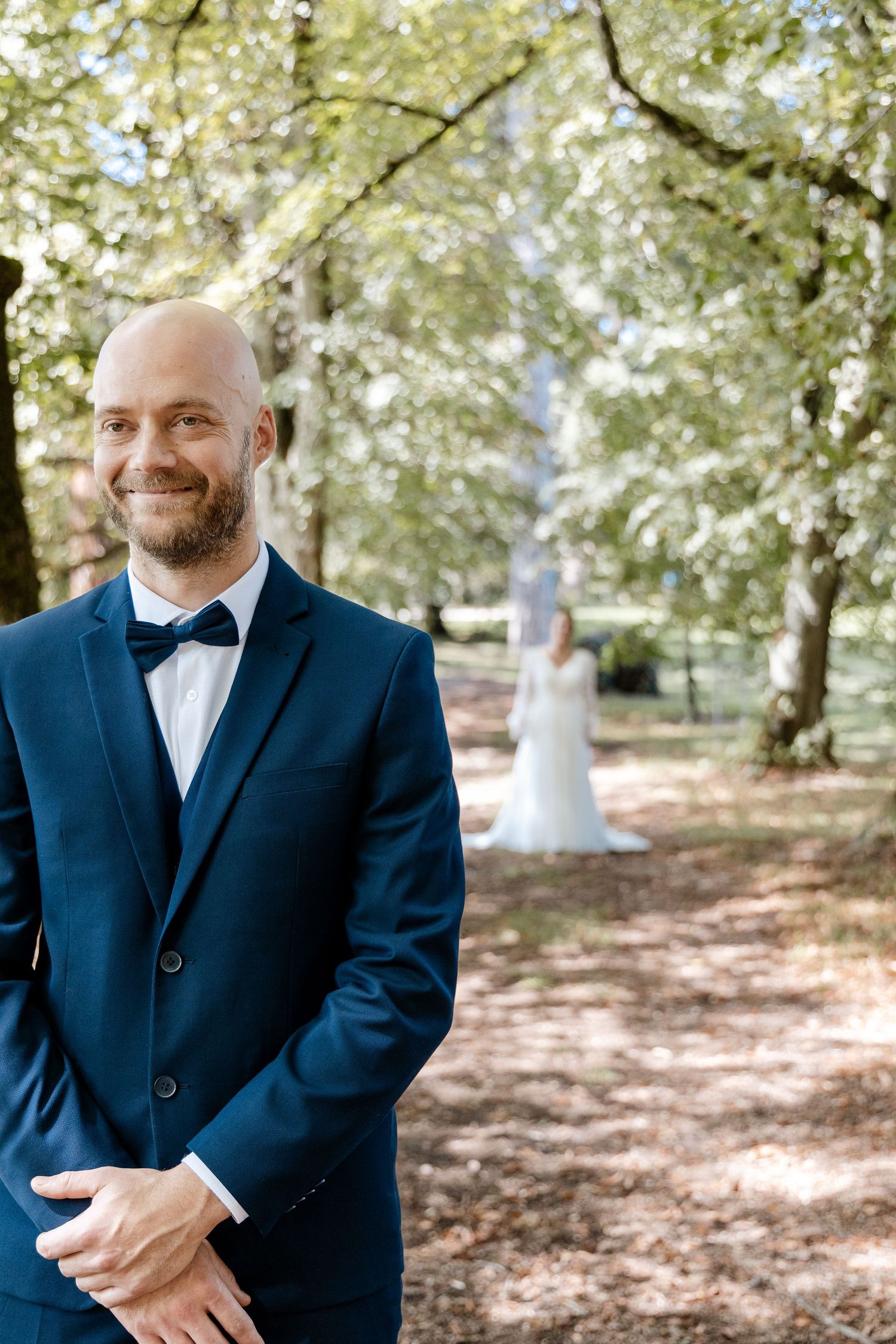Smiling groom in navy suit with bow tie stands in sunlit forest path, bride in white gown visible in background.