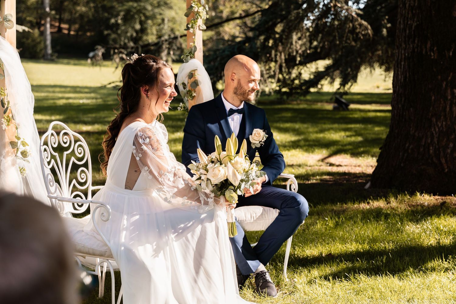 Bride and groom sit together smiling during their sunny outdoor wedding ceremony in a lush garden.