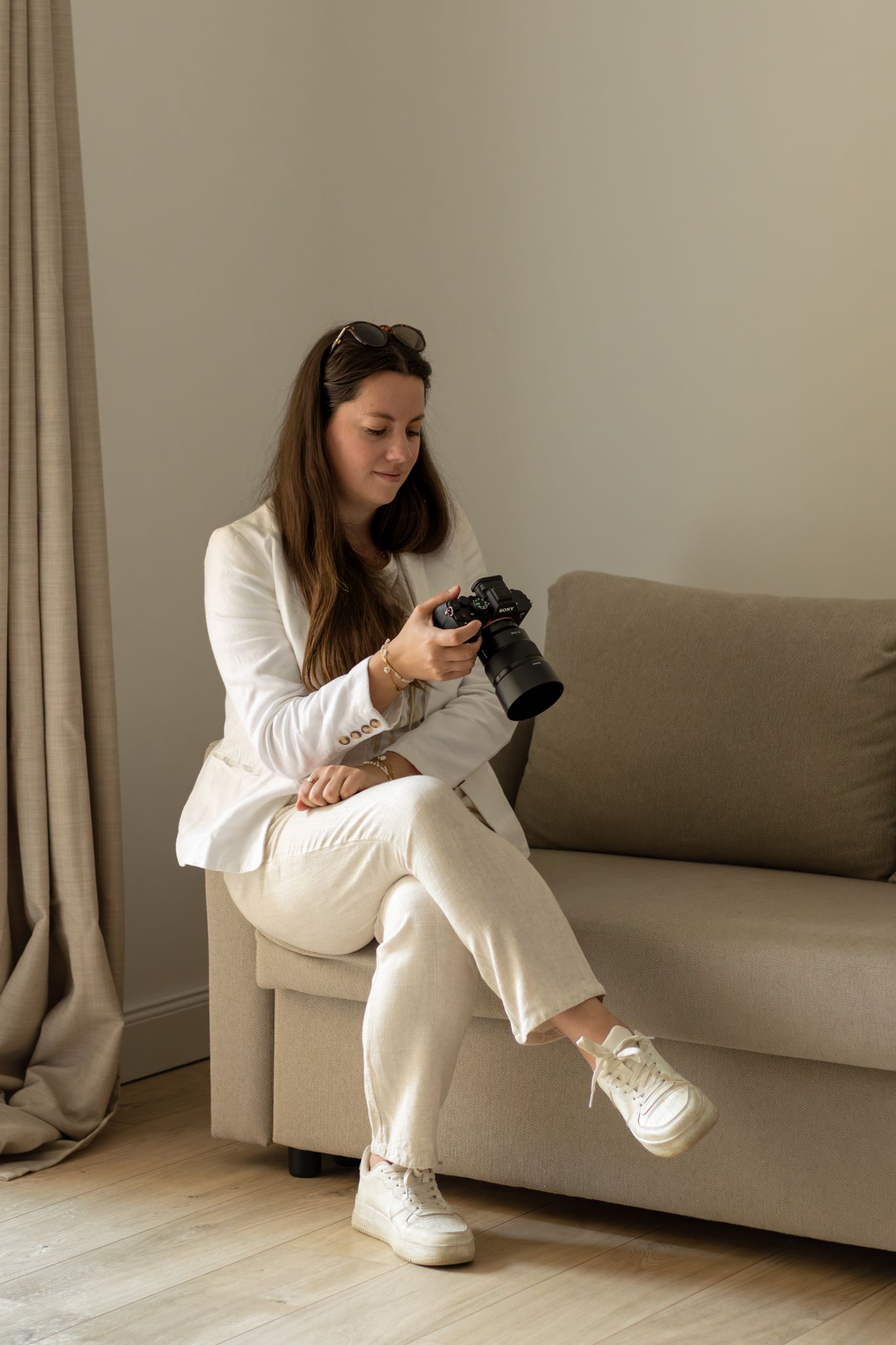 Woman in white suit sitting on a sofa reviewing photos on a professional camera indoors.