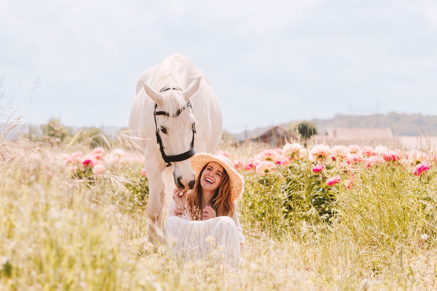 A woman in white sits laughing under a white horse in a blooming flower field.