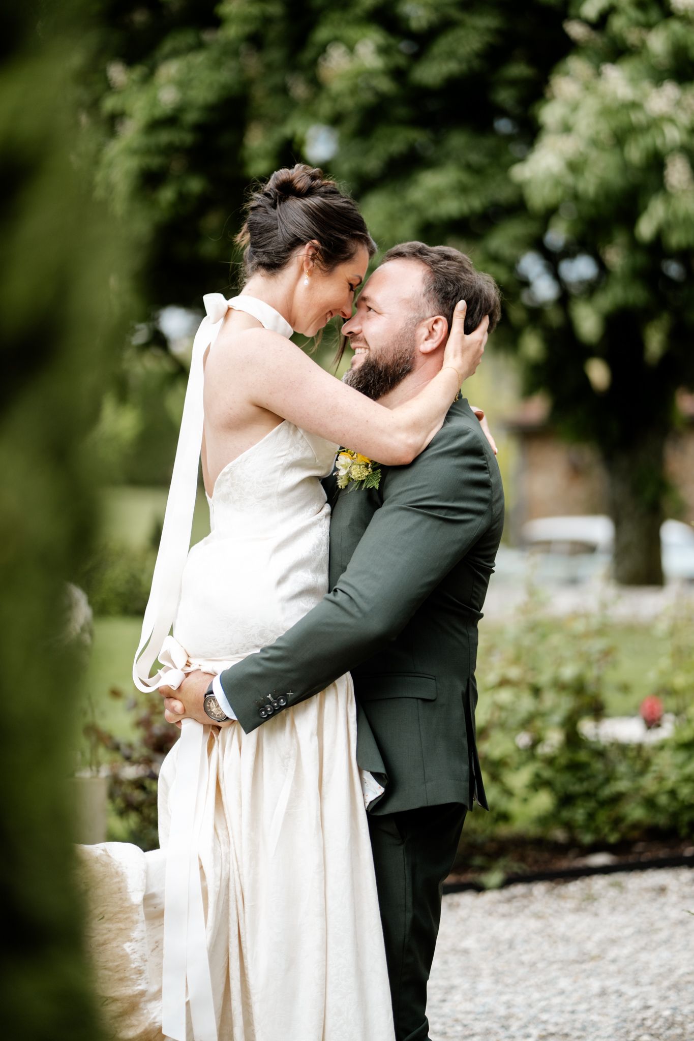 Groom in dark green suit lifting bride in white halter gown for romantic embrace in garden.