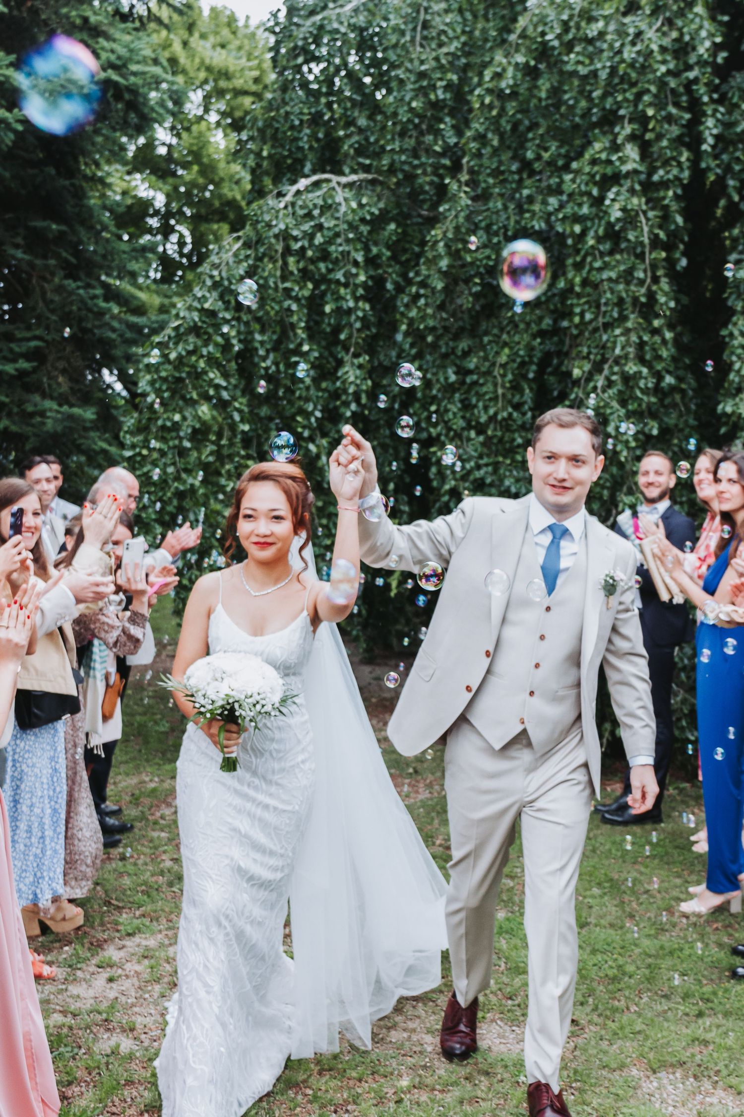 Newlyweds walk joyfully through a tunnel of guests blowing bubbles during outdoor wedding celebration.