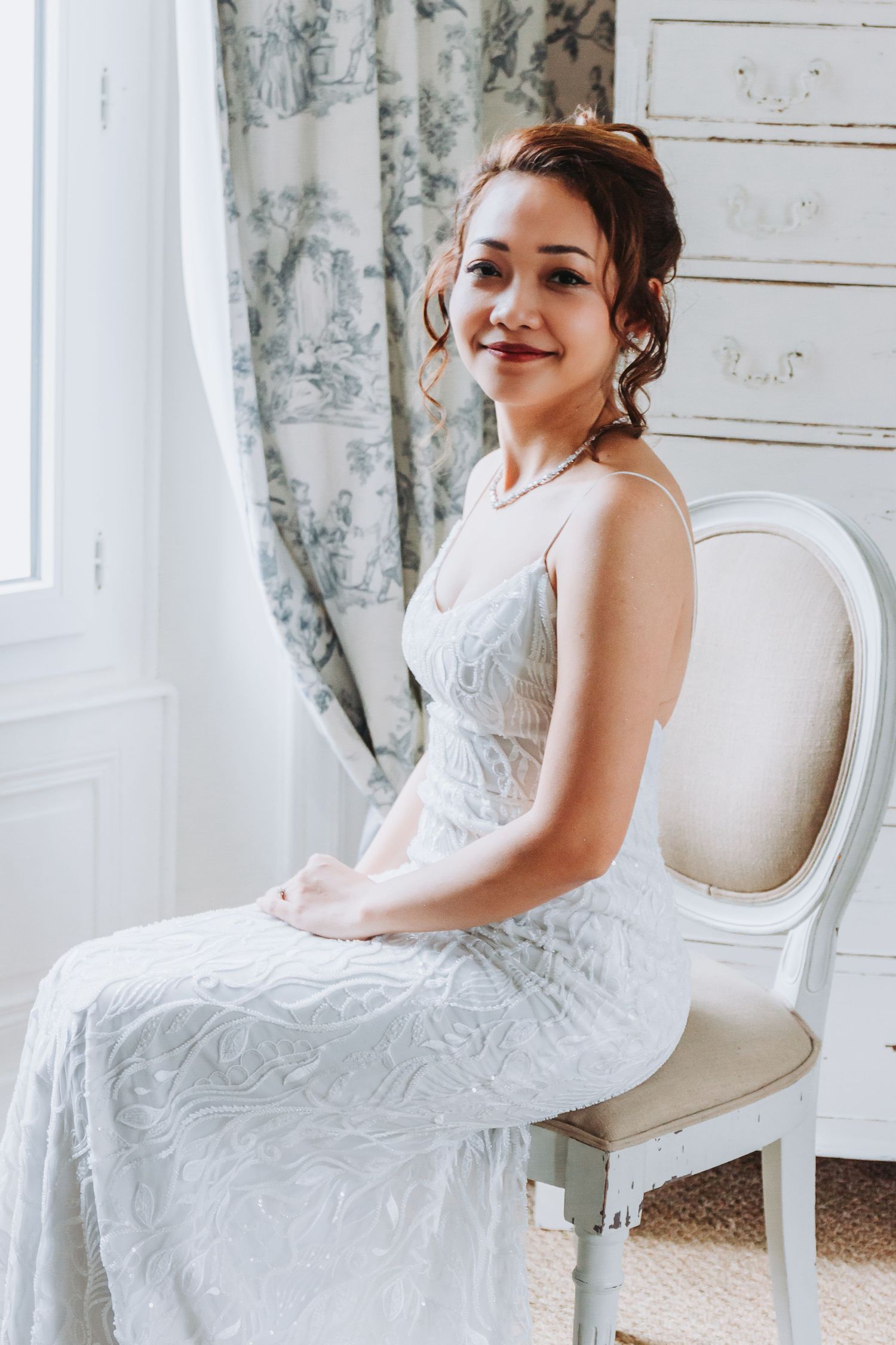 Smiling Asian bride in a white lace wedding dress seated on a French chair by a toile curtain window.