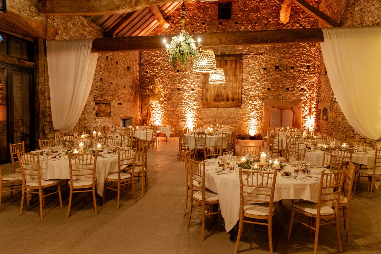 Rustic stone barn wedding reception with round tables, gold chairs, candles, and warm amber lighting.