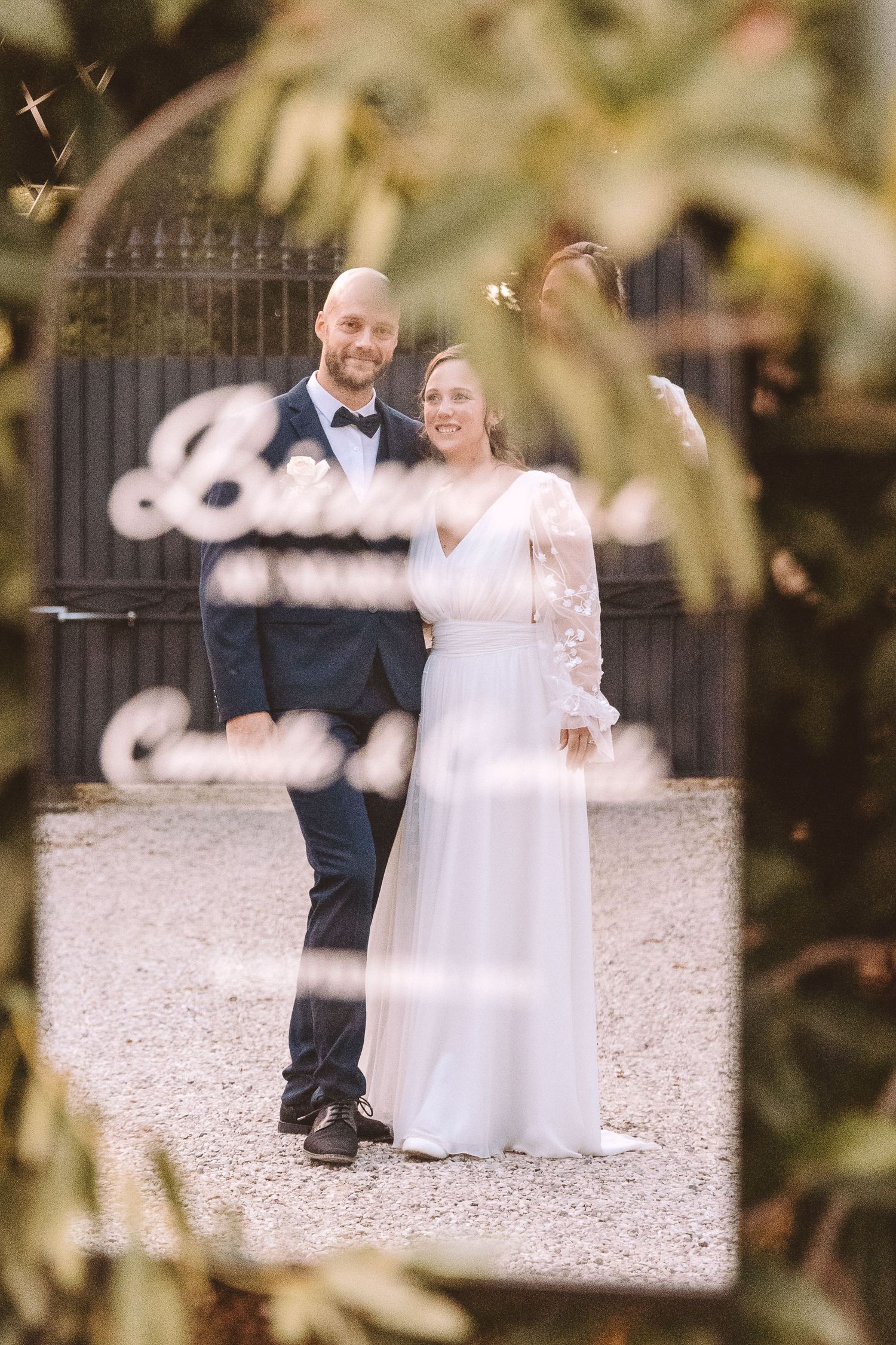 Bride and groom smiling together at their wedding venue, framed by lush green foliage.