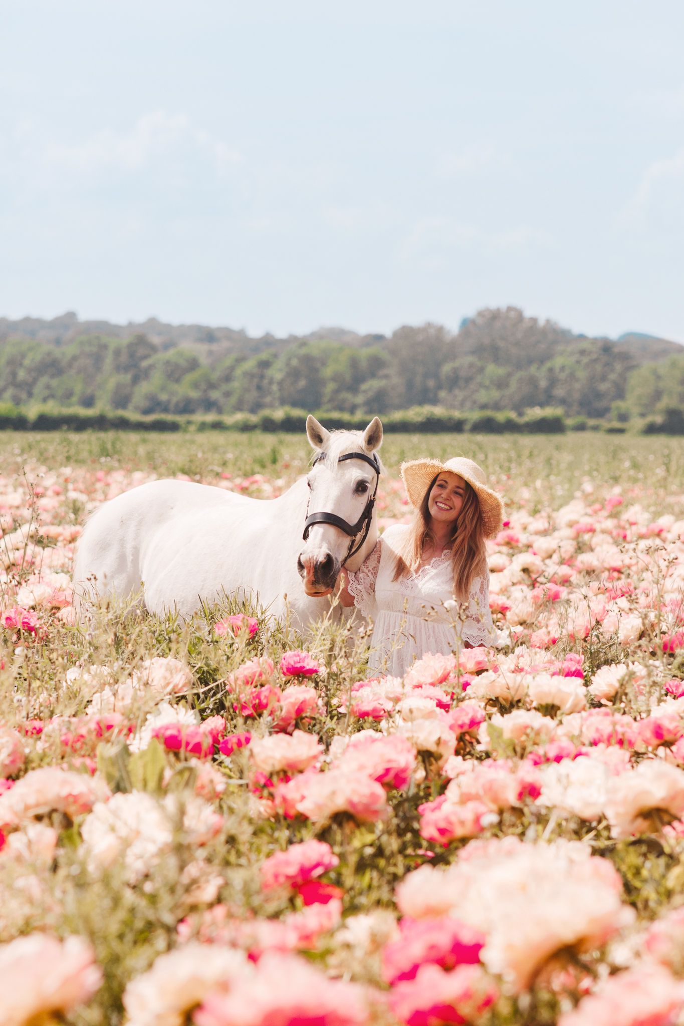 Woman in white dress and straw hat standing with white horse in pink flower field.