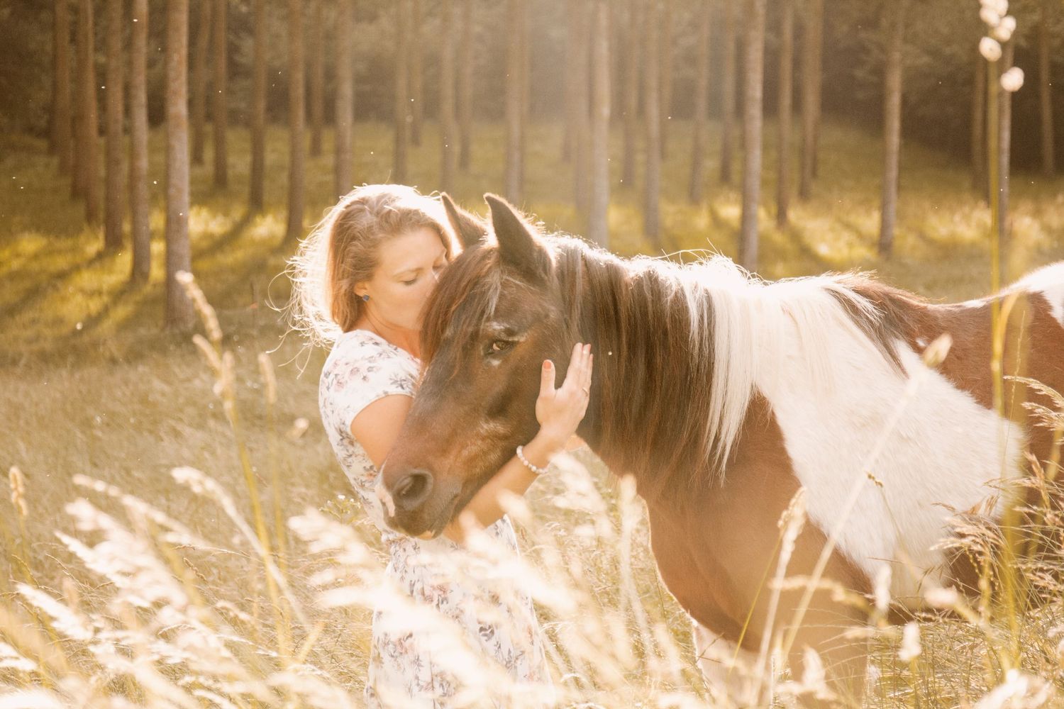Young woman tenderly embracing a paint horse in a sunlit forest clearing.