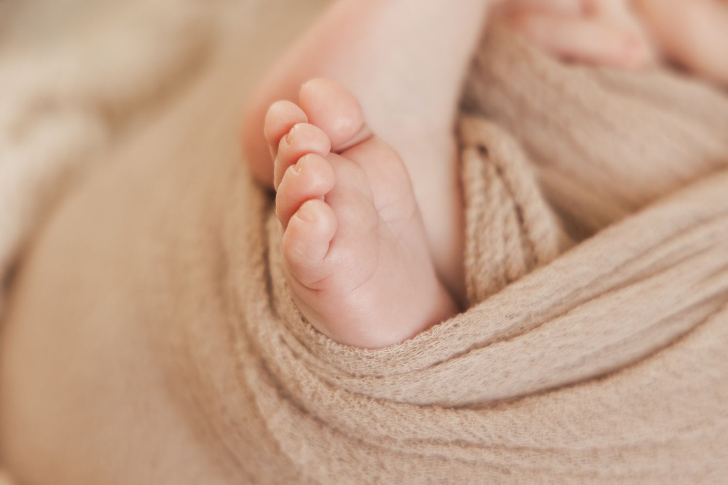 Close-up of tiny newborn baby feet wrapped in soft beige swaddle blanket.