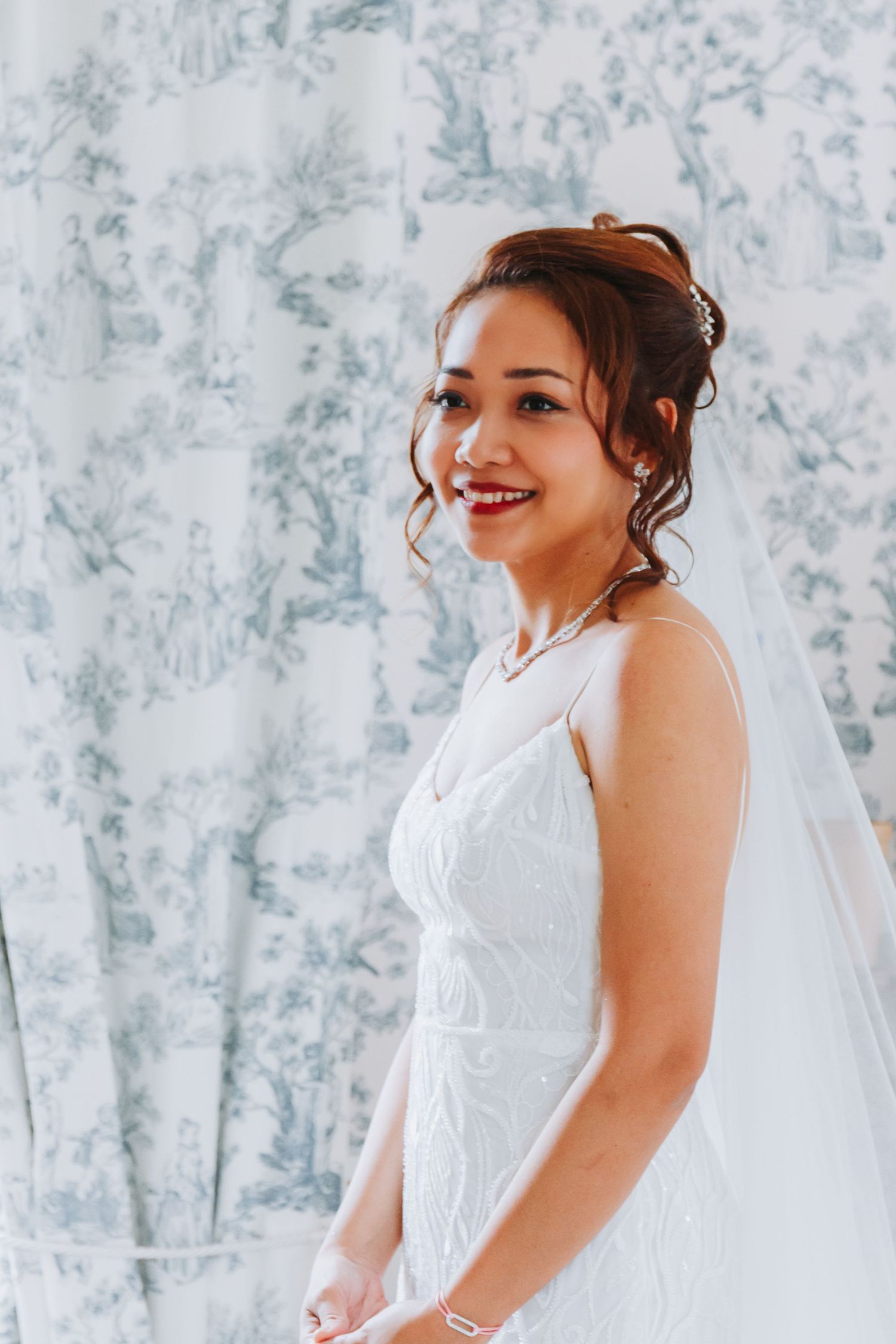 Radiant Asian bride smiling in a white lace gown with veil, standing before blue toile de jouy curtains.
