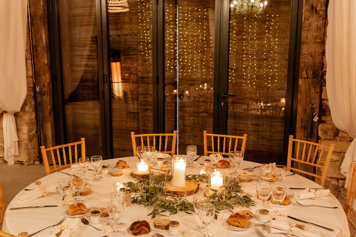 Elegant round wedding table with greenery centerpiece, candles, and fairy light backdrop in rustic venue.