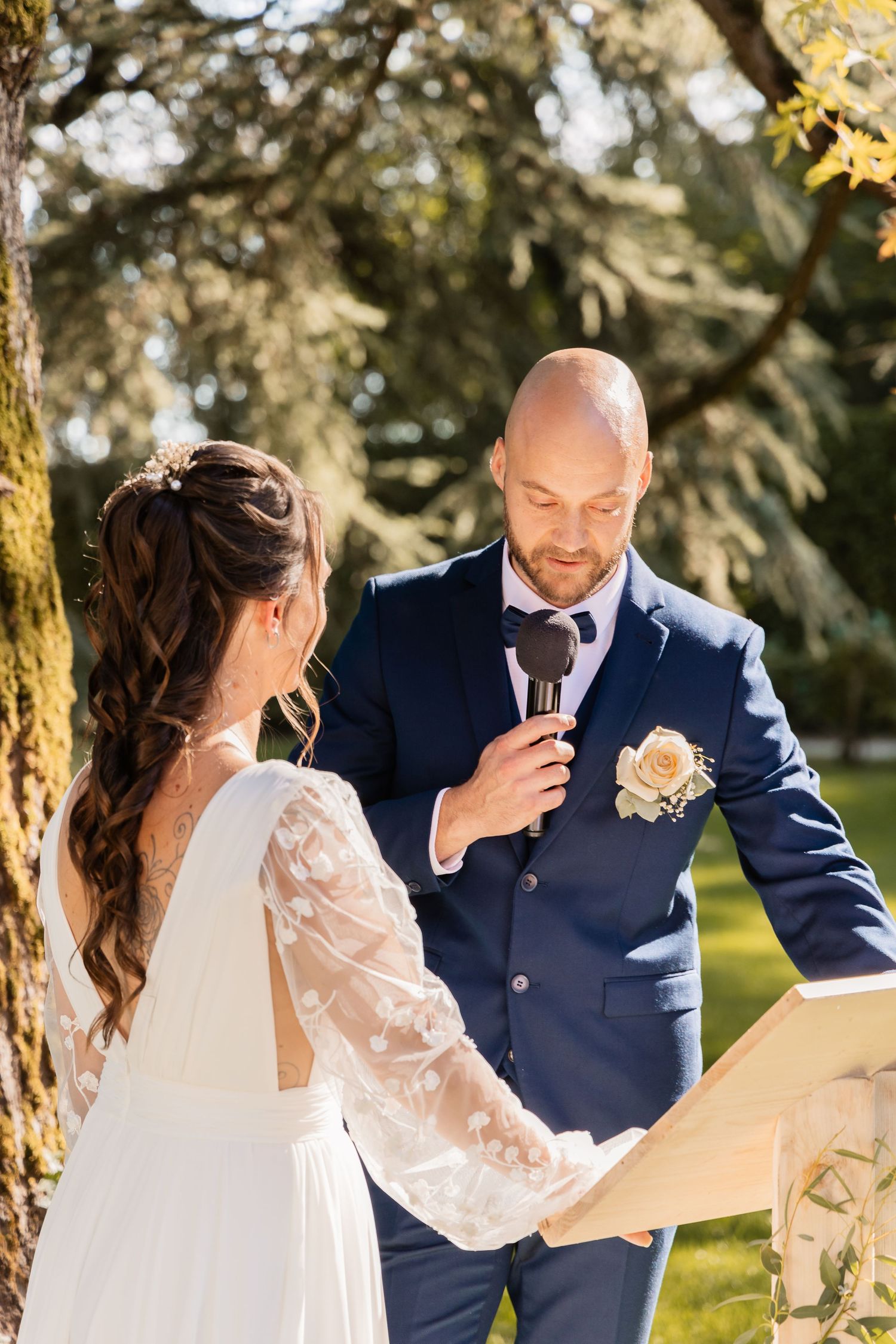 Groom reads wedding vows into a microphone while holding the bride's hand at an outdoor ceremony.