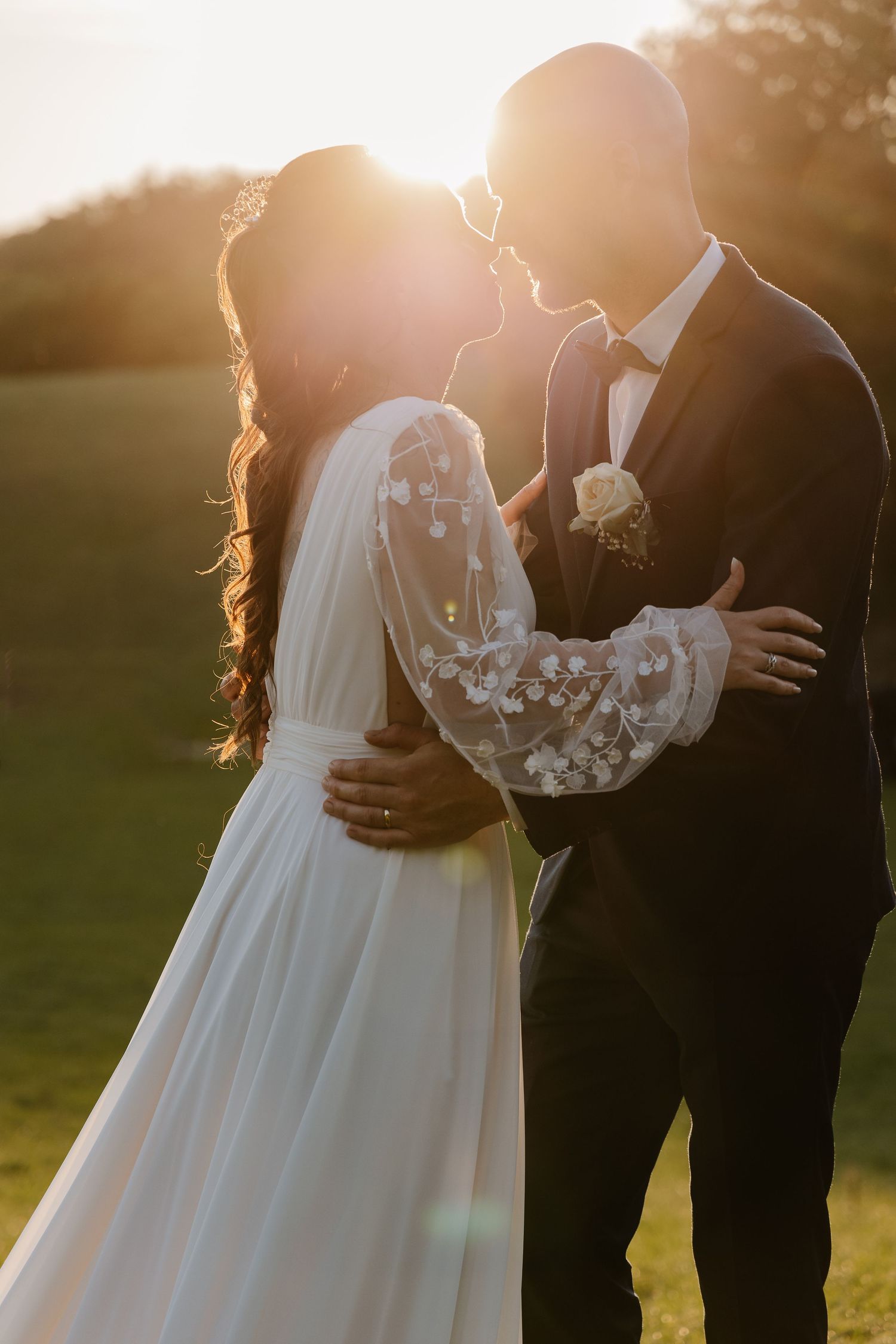 Bride and groom share a kiss at golden sunset, bride wearing floral lace long-sleeve wedding gown outdoors.
