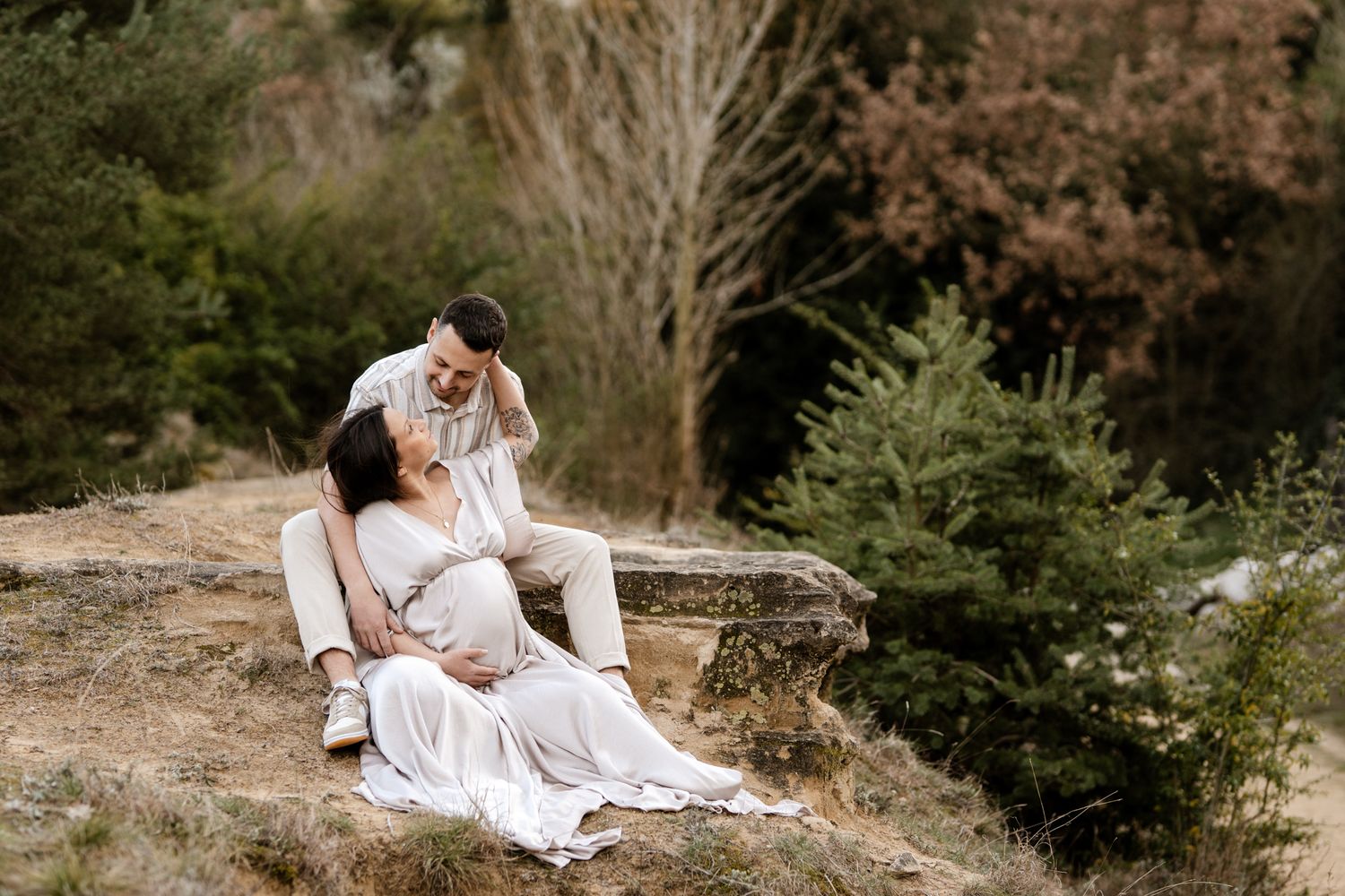 Séance photo grossesse en Isère : un couple se détend sur des rochers, le futur papa sourit à la future maman en robe nacrée.